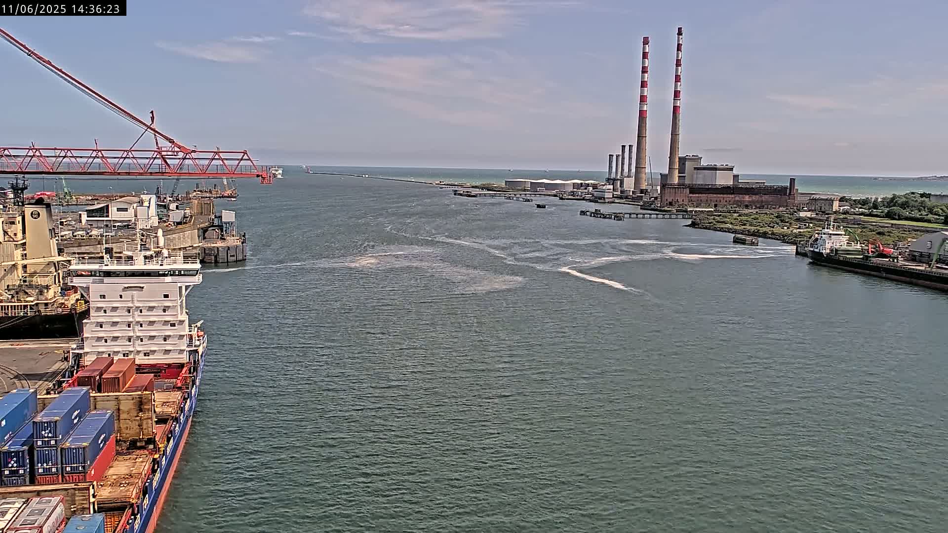 A container ship is docked near a port with two large industrial smokestacks in the background on a partly sunny day.