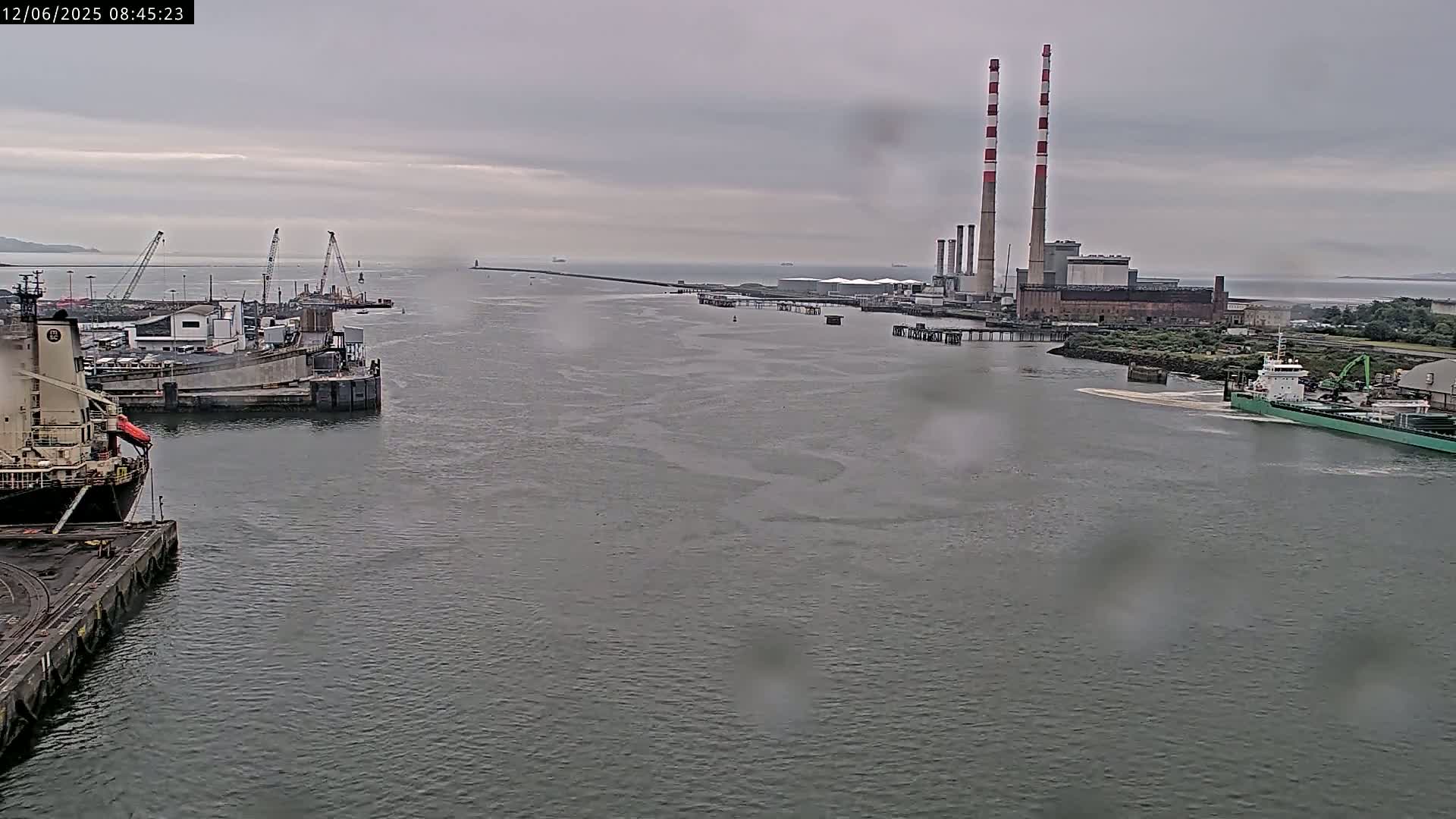 A wide shot of a harbor on an overcast day shows several docked ships, cranes, and a power plant with tall smokestacks in the distance, all under a rainy sky.