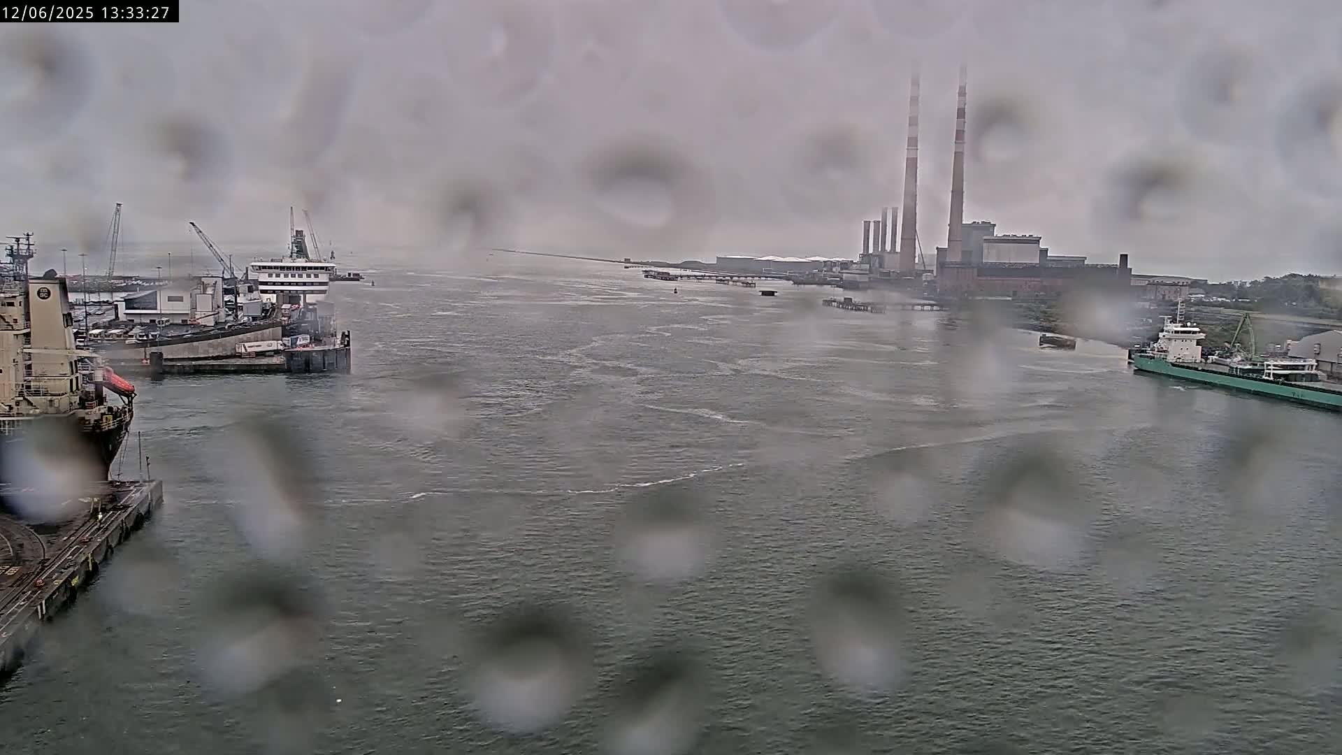 A rain-streaked view of a harbor shows several ships docked, with a power plant and other industrial buildings visible in the background.