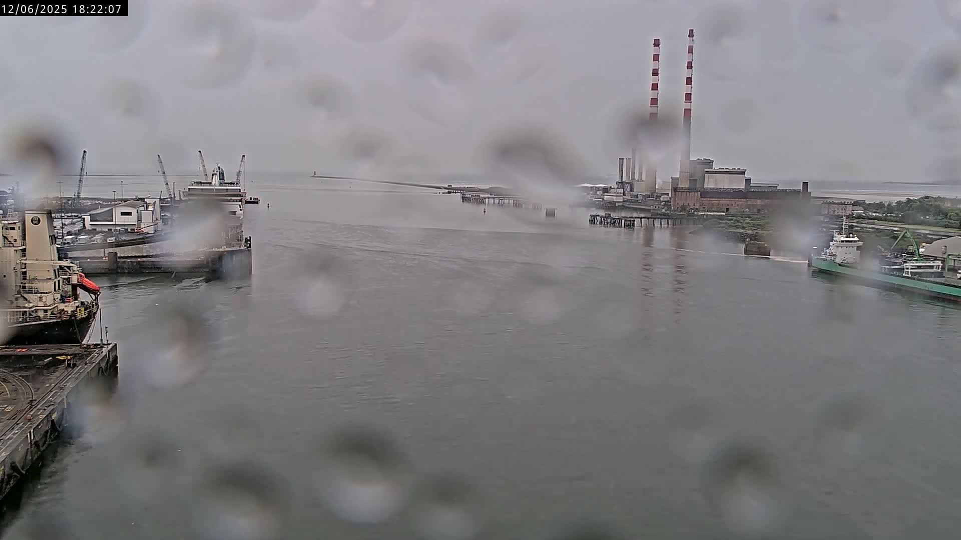 Rain-streaked view of a harbor with several ships docked, a power plant with tall smokestacks in the background, and a body of water extending to a distant shoreline.