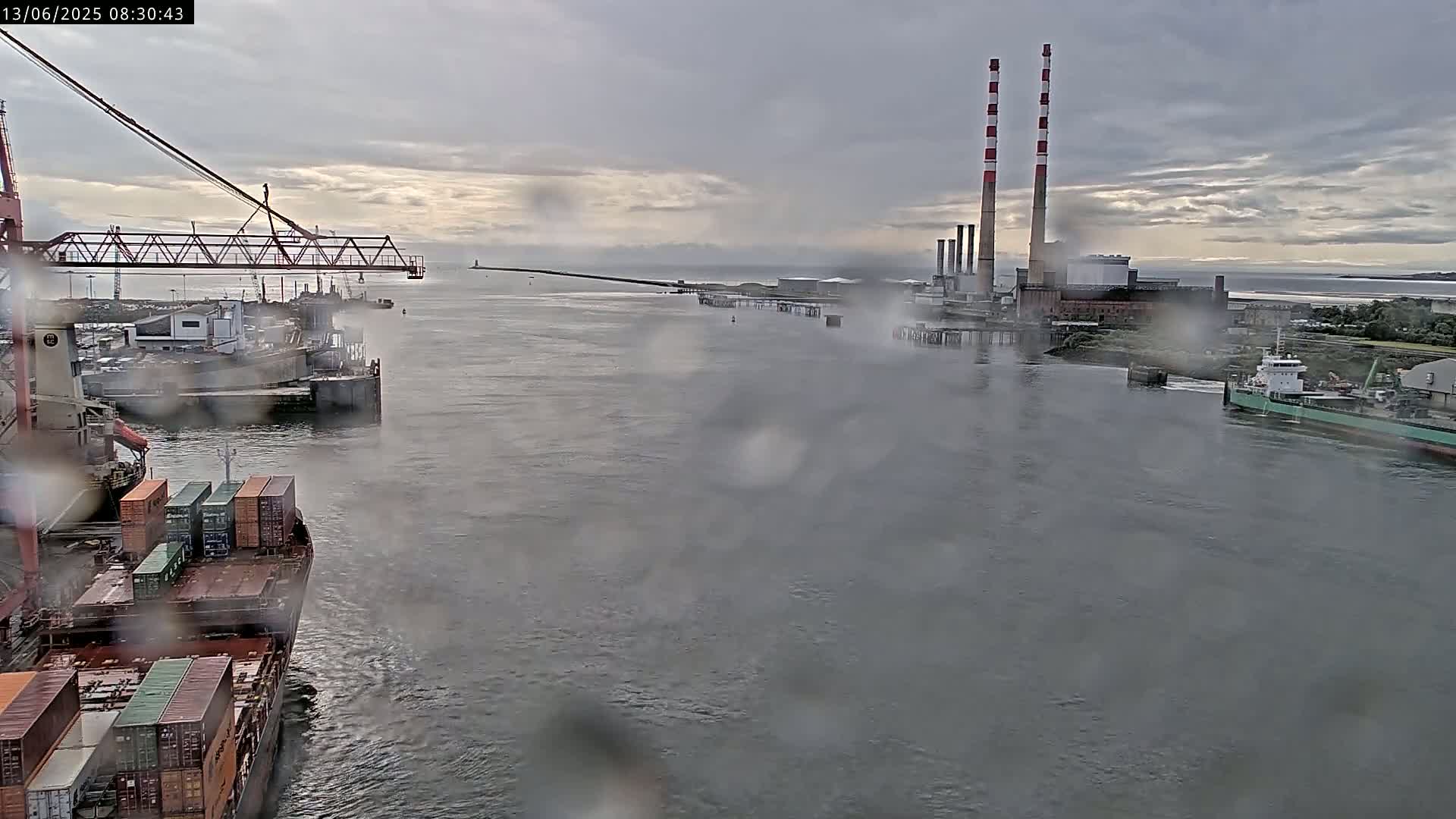 A rainy view of a harbor shows a container ship, a large industrial plant with tall smokestacks, and other vessels on the water.