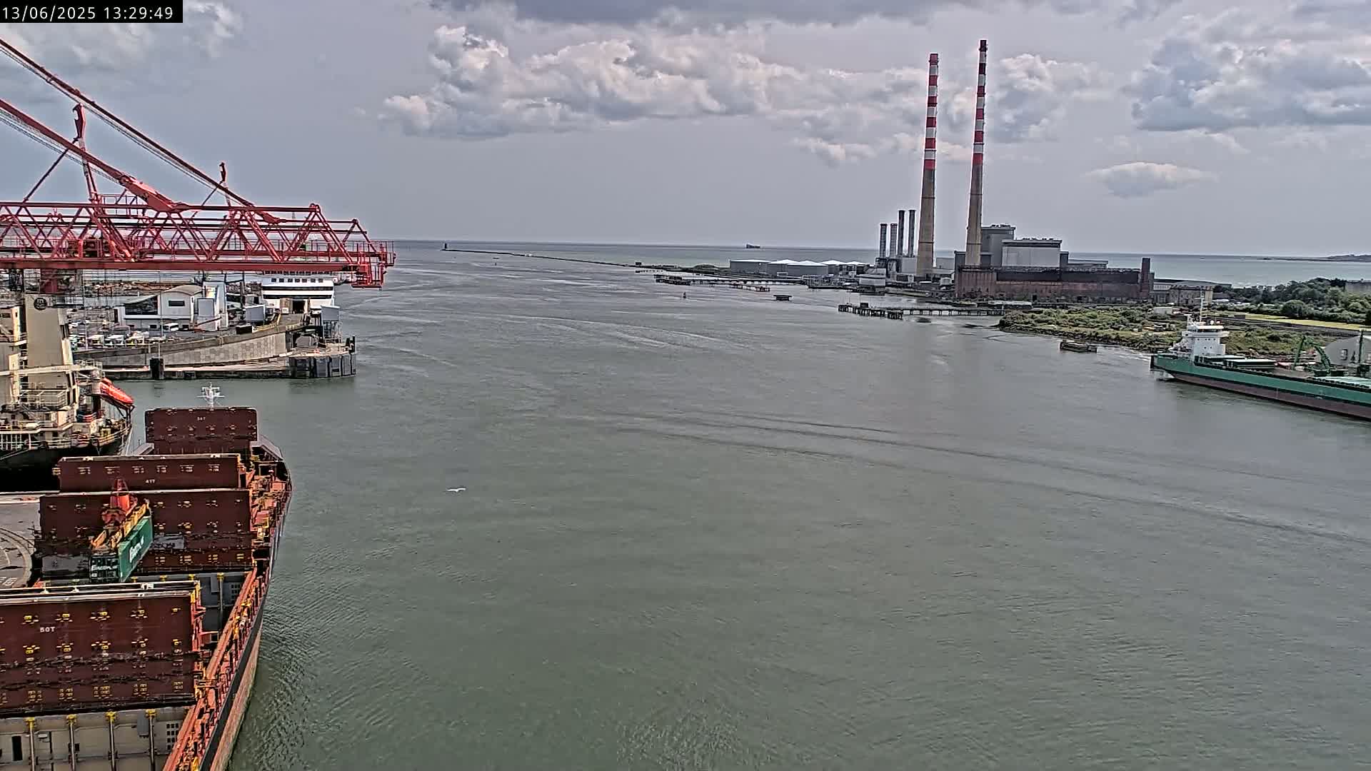 A container ship is docked at a port under partly cloudy skies, with a power plant and other vessels visible in the distance.