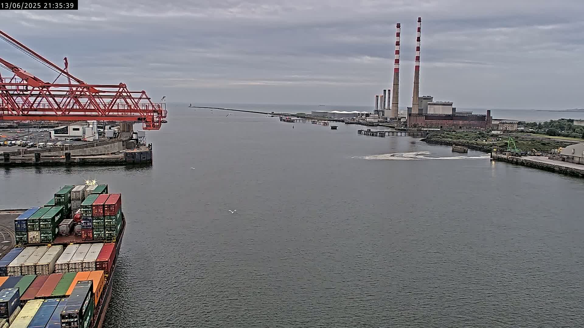 A container ship sits in a harbor next to an orange gantry crane, with a large industrial plant and tall smokestacks visible in the distance under an overcast sky.