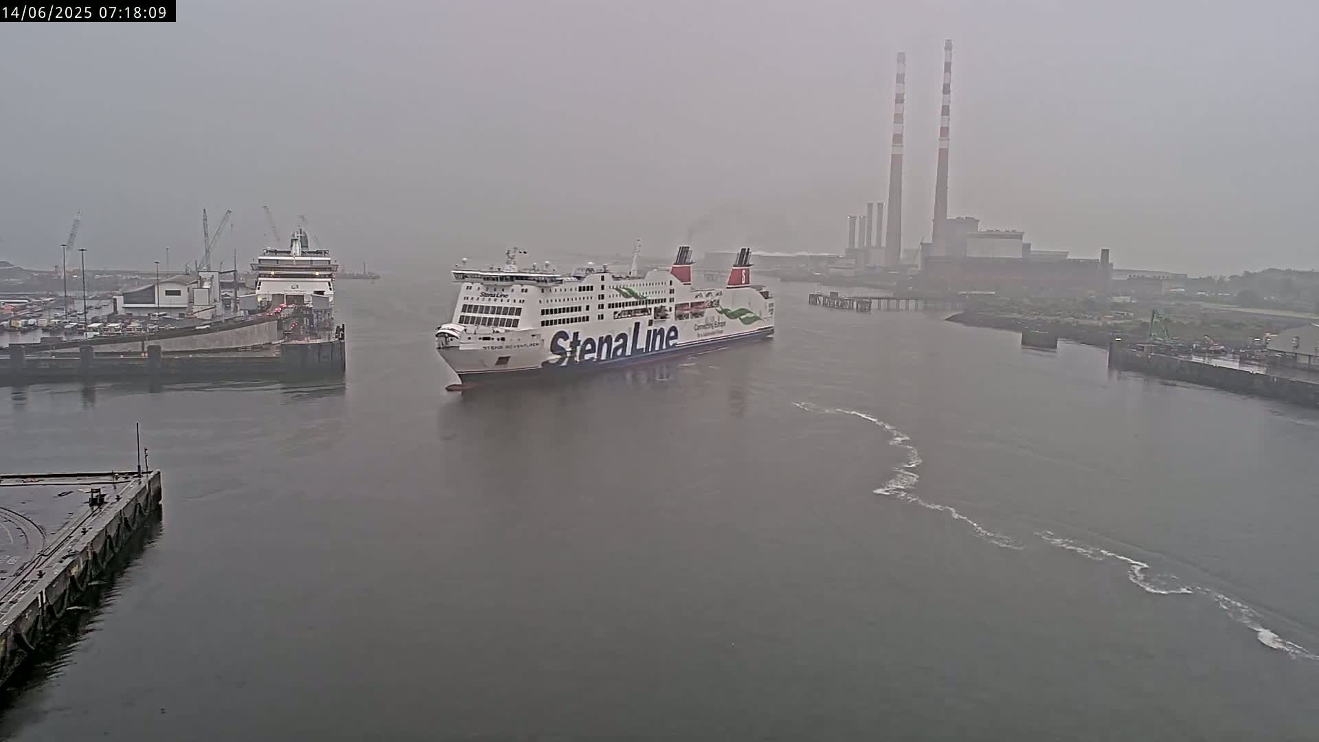 A container ship sits in a harbor next to an orange gantry crane, with a large industrial plant and tall smokestacks visible in the distance under an overcast sky.