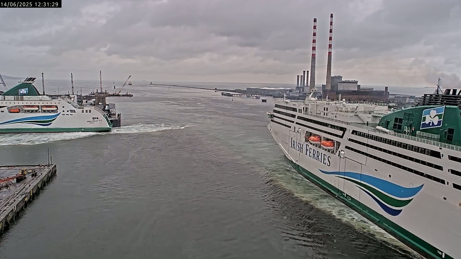 Two ferries are in a harbor on an overcast day, one docked and one approaching a dock near a power plant with tall smokestacks.