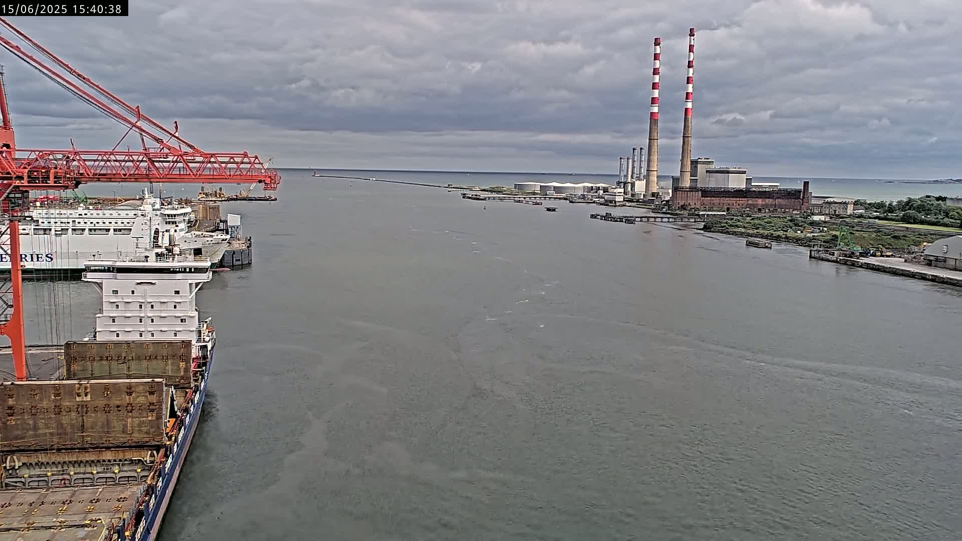 A large cargo ship is docked near a ferry and a large industrial facility with tall smokestacks under a cloudy sky.