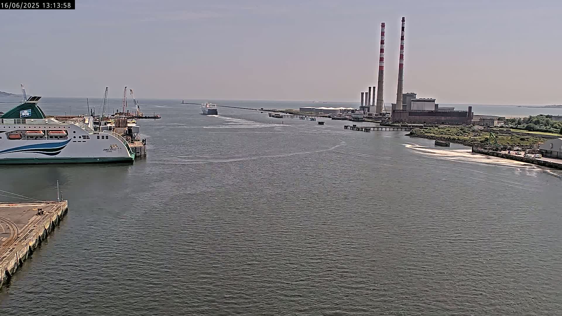 A ferry is docked at a port near a power plant with two tall smokestacks, on a calm, sunny day.