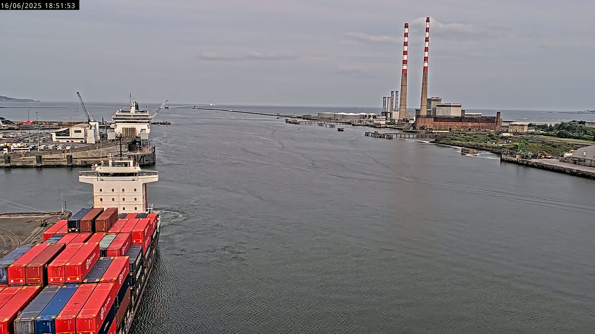 A container ship sails past a docked ferry in a harbor under overcast skies, with a large industrial plant featuring tall smokestacks visible in the distance.