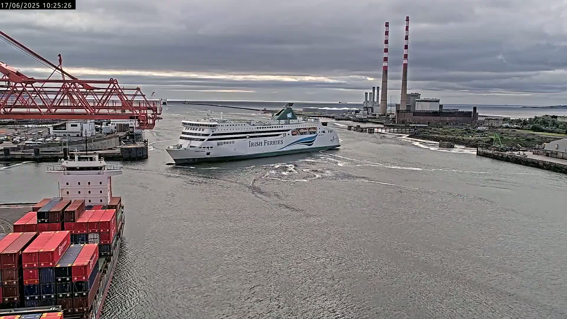 A ferry passes a container ship in a harbor under an overcast sky, with industrial structures and tall smokestacks visible in the background.