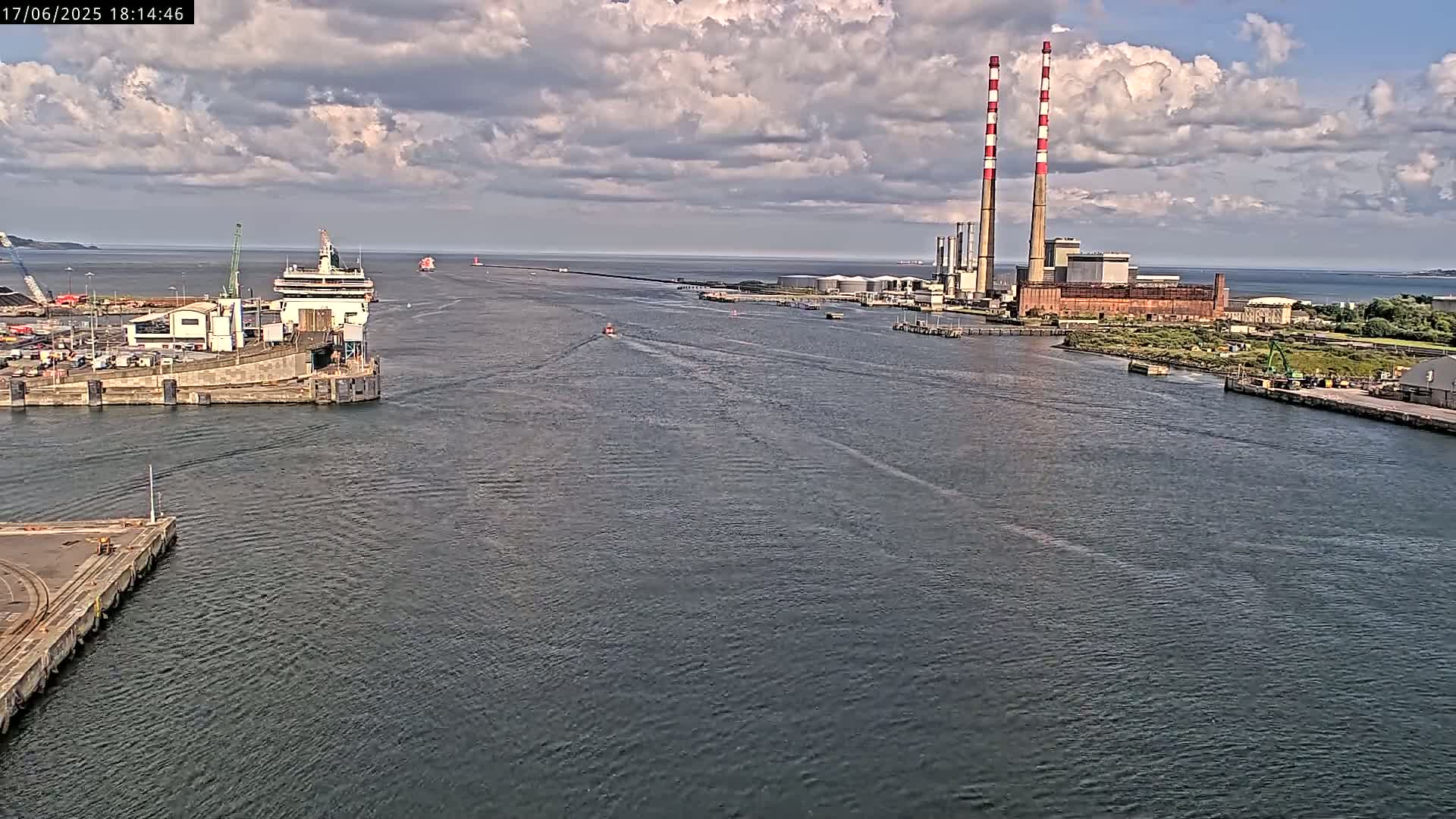 A large ship is docked at a harbor under a partly cloudy sky, with two tall smokestacks visible in the distance.