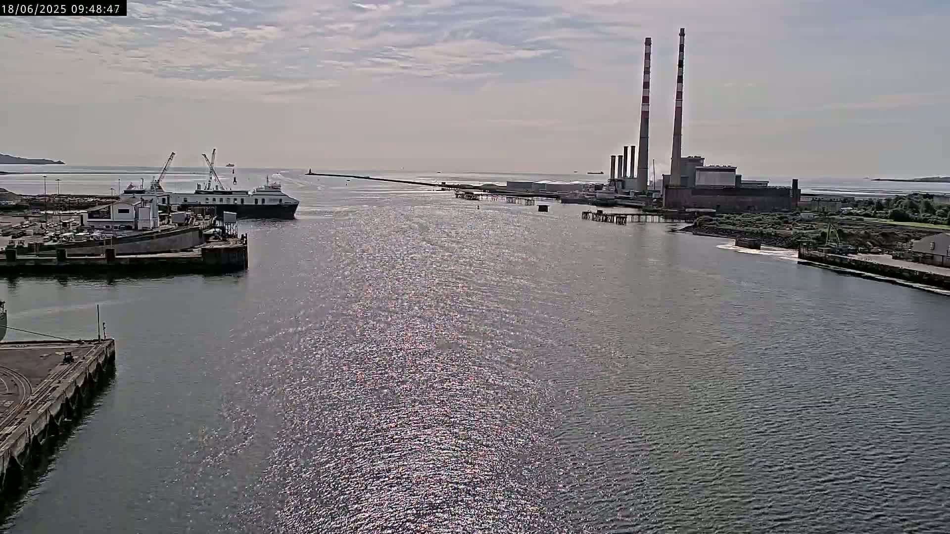 A ferry is docked at a harbor near a power plant under a partly cloudy sky.