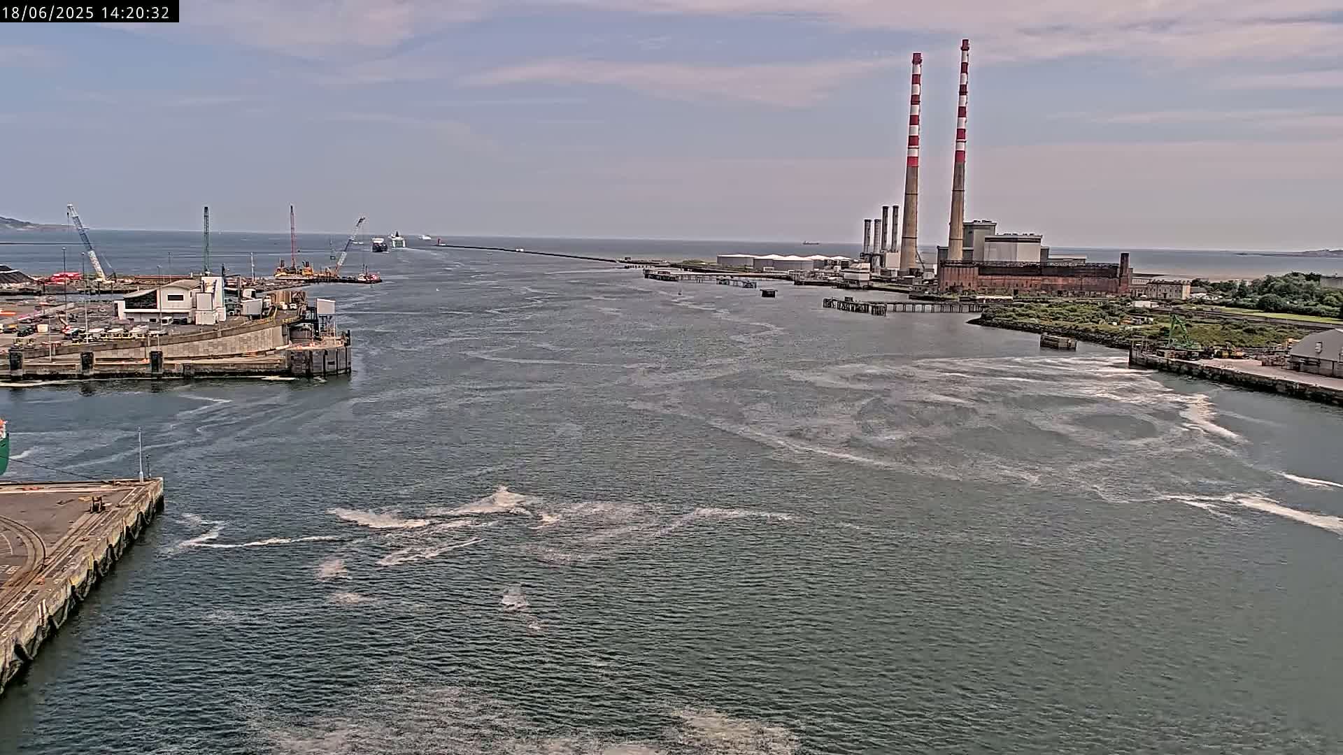 A wide view of a harbor on a partly cloudy day shows a large industrial plant with two tall smokestacks on the right, a ship in the distance, and various docks and equipment in the foreground.