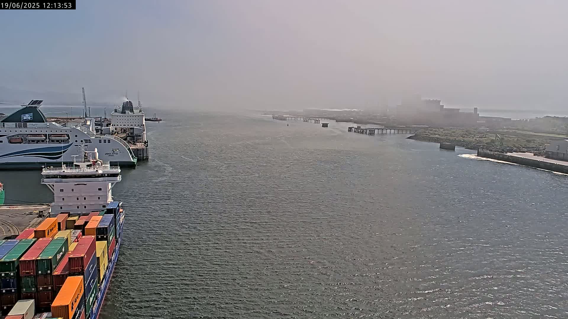 A container ship is docked next to two ferries in a hazy harbor.