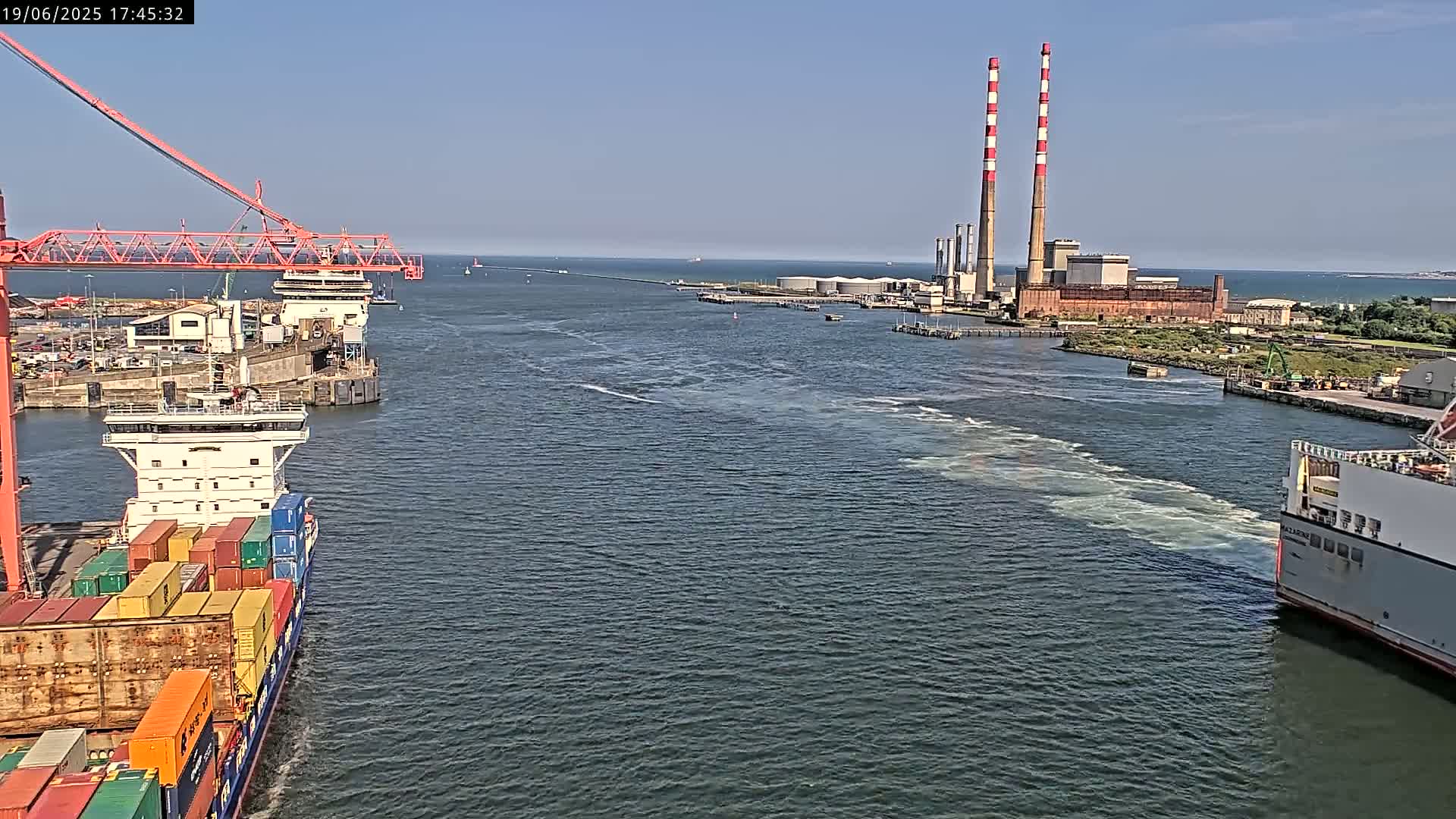 A container ship and a ferry are in a harbor on a sunny day, with two tall smokestacks visible in the distance.