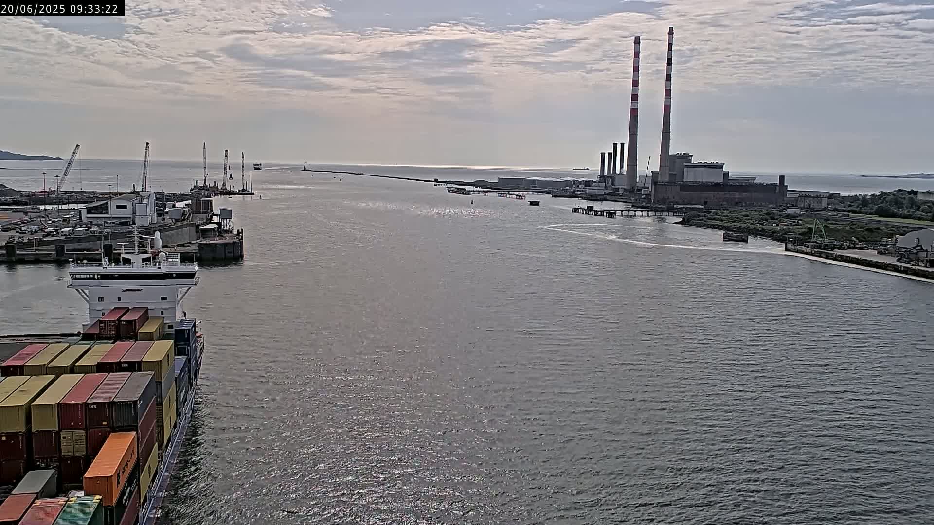 A container ship sails through a harbor toward a large industrial plant with tall smokestacks under a partly cloudy sky.