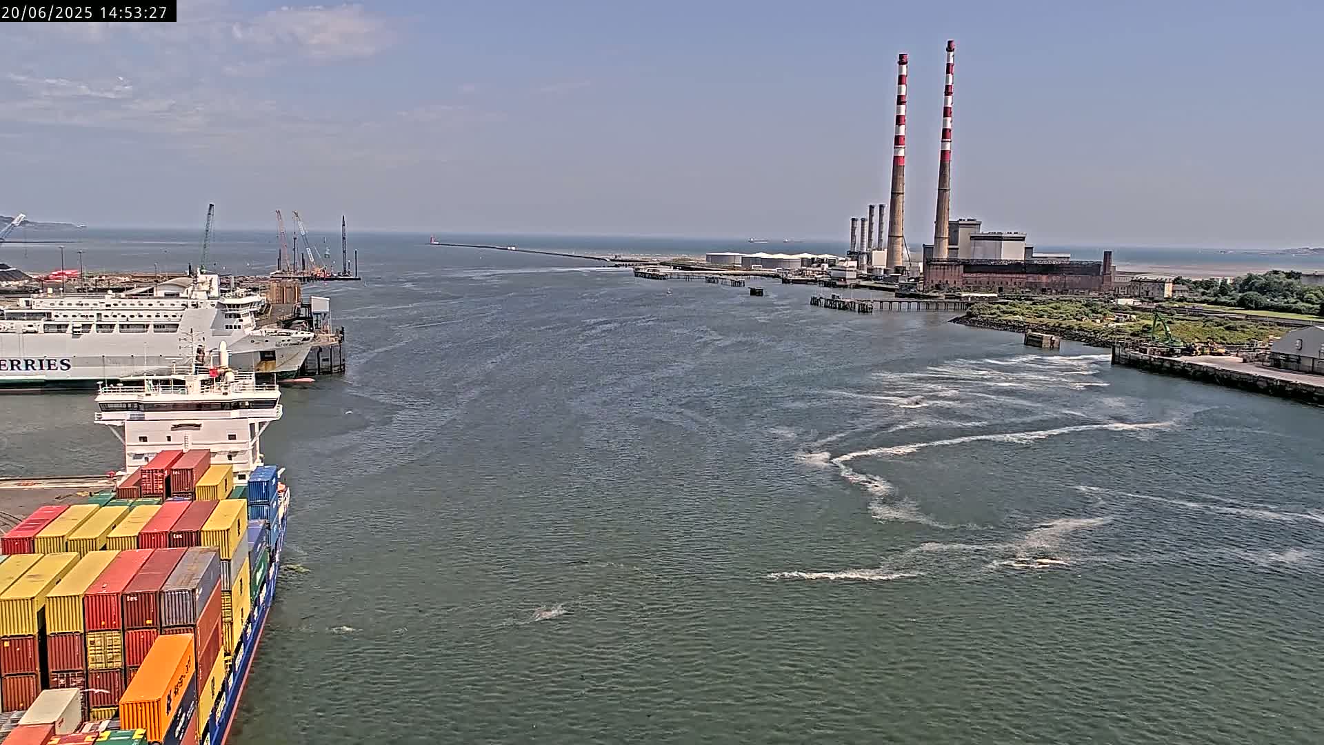 A container ship and a ferry are docked near a power plant with tall red and white smokestacks on a sunny day, with choppy water visible in the harbor.
