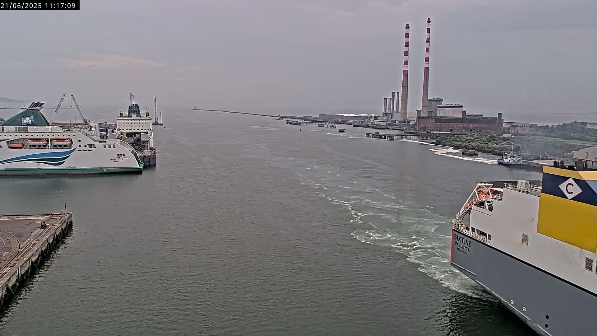 Two ferries are in a harbor on an overcast day, one docked and another passing by a power plant with tall smokestacks.