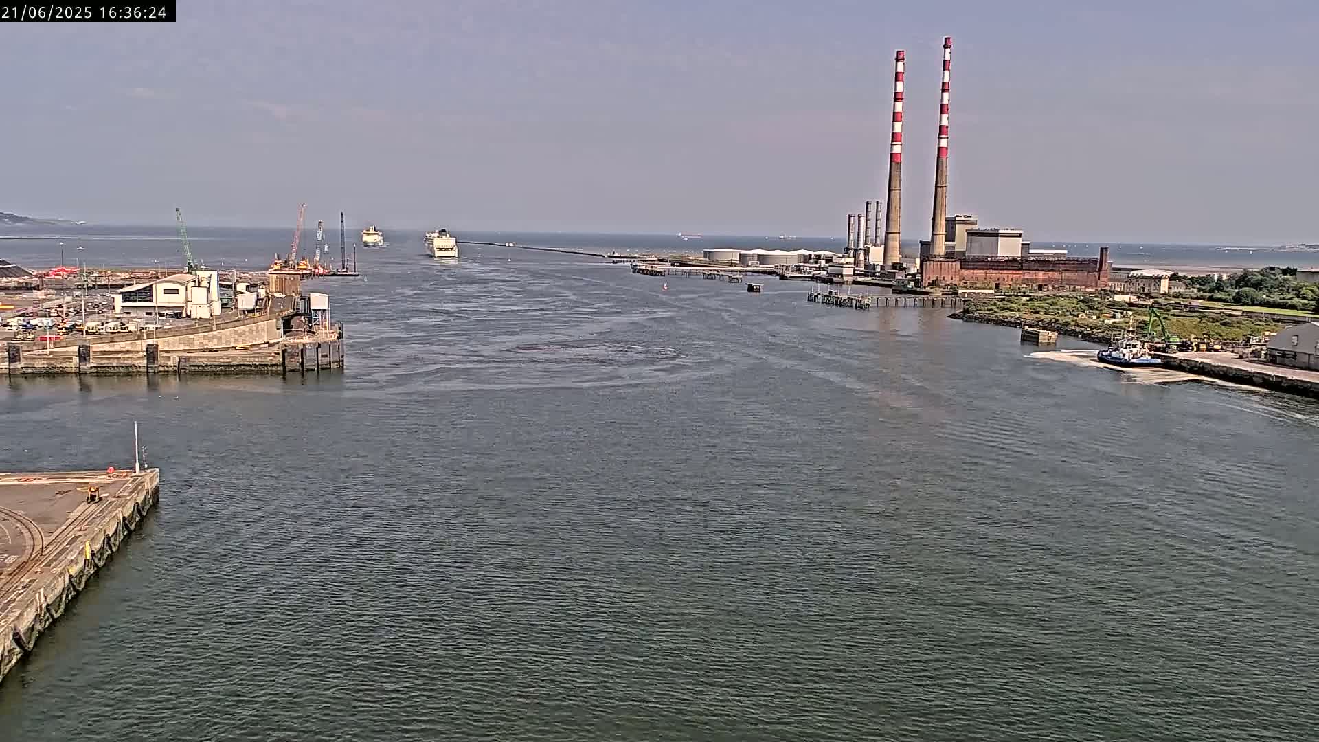 A wide shot shows a calm body of water with several ships near a harbor and industrial buildings with tall smokestacks on a sunny day.