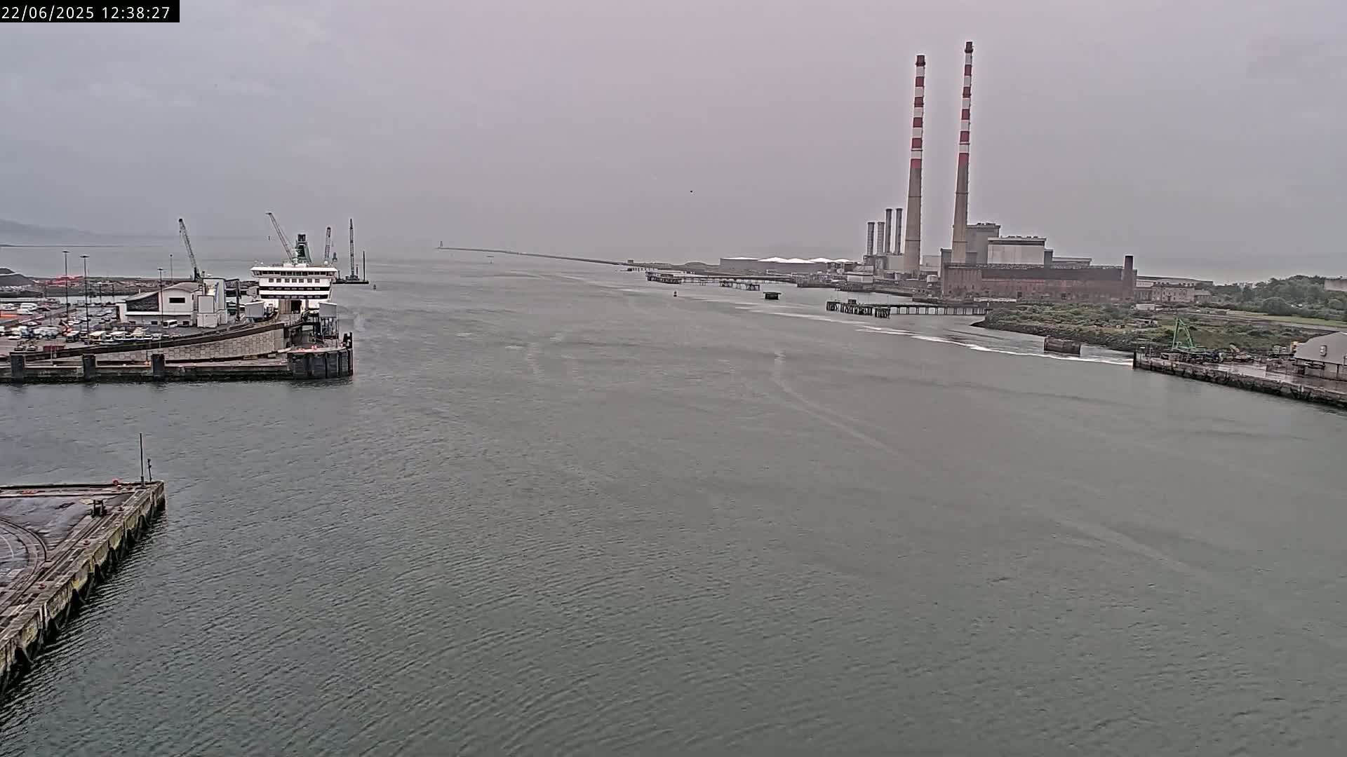 A ferry is docked at a port on an overcast day, with a power plant visible in the background across a calm body of water.