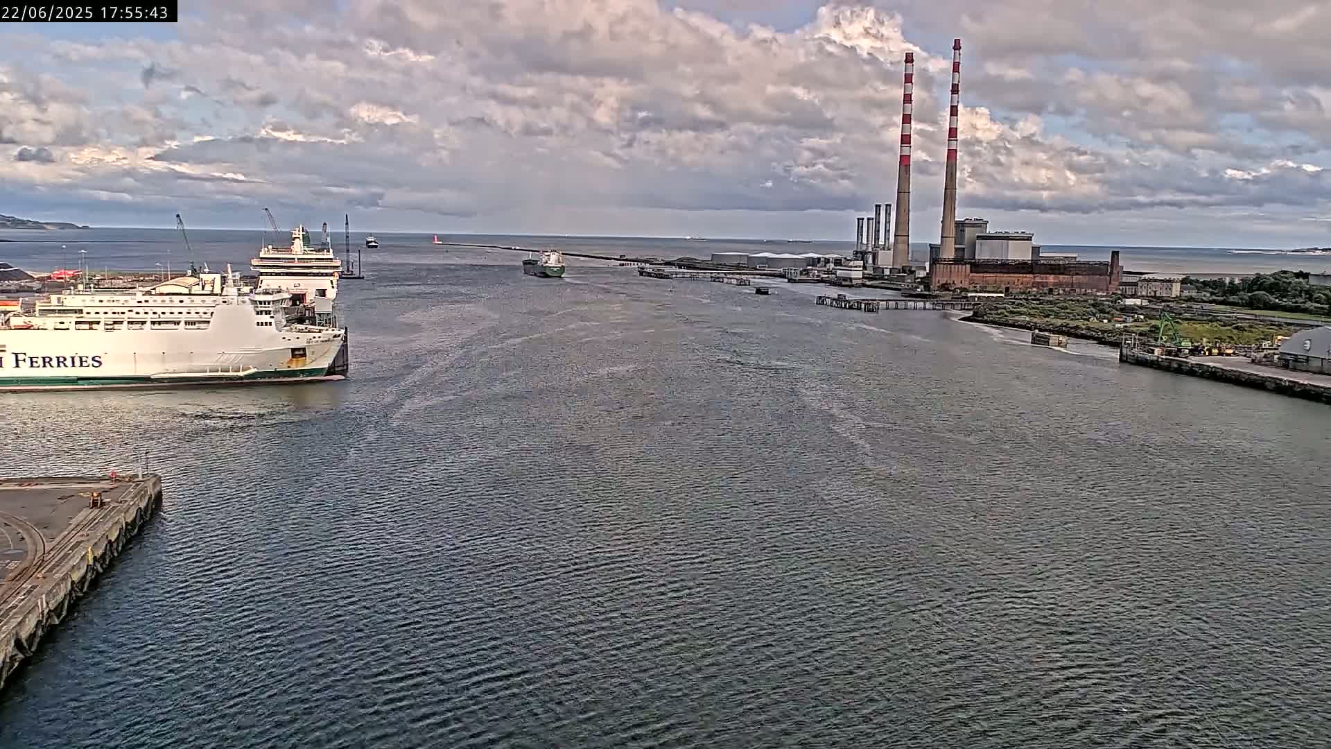 Two ferries are docked near a power plant with two tall smokestacks under a partly cloudy sky.