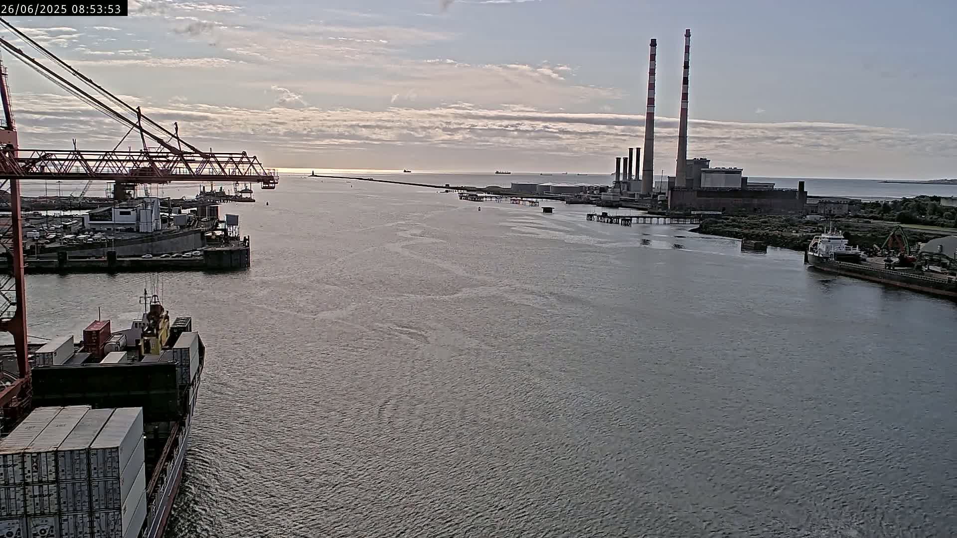 A container ship sails past a port with a large industrial plant and tall smokestacks visible in the distance under a partly cloudy sky.