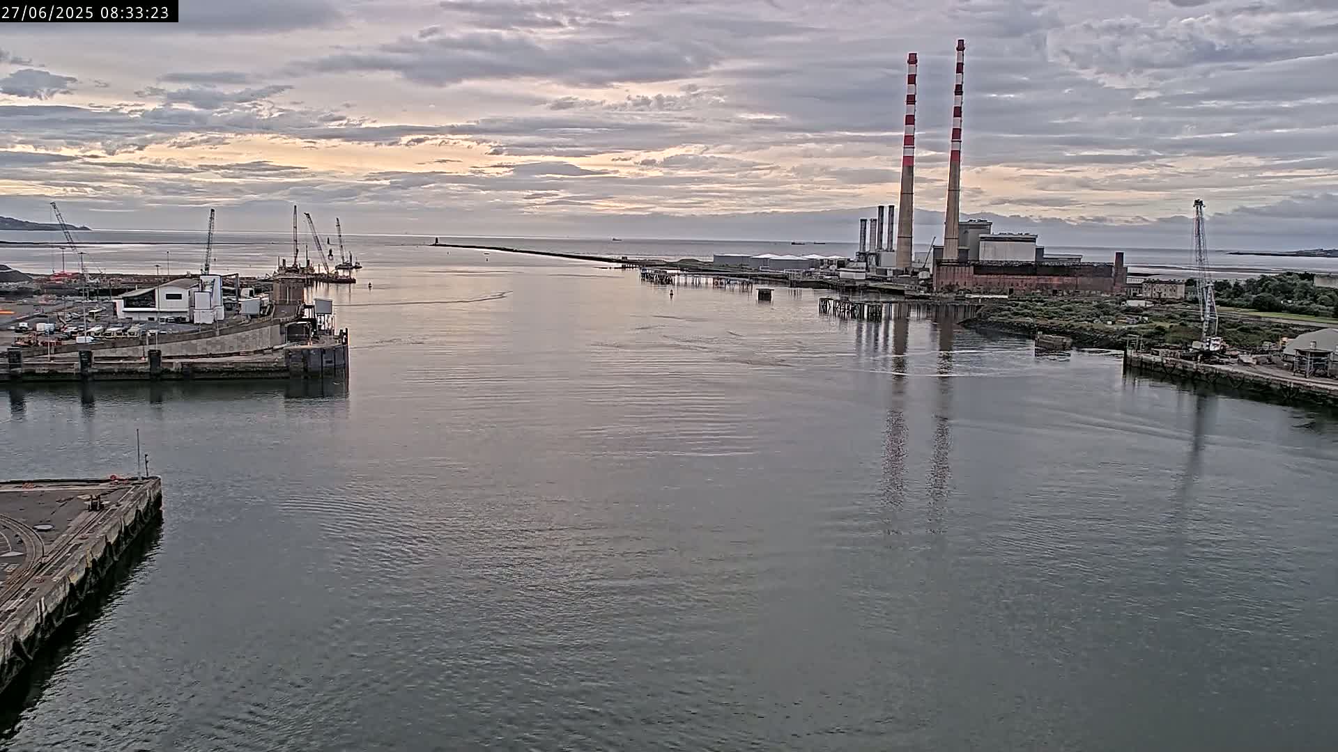 A calm body of water shows a harbor with industrial buildings, cranes, and two tall smokestacks under a cloudy sky.