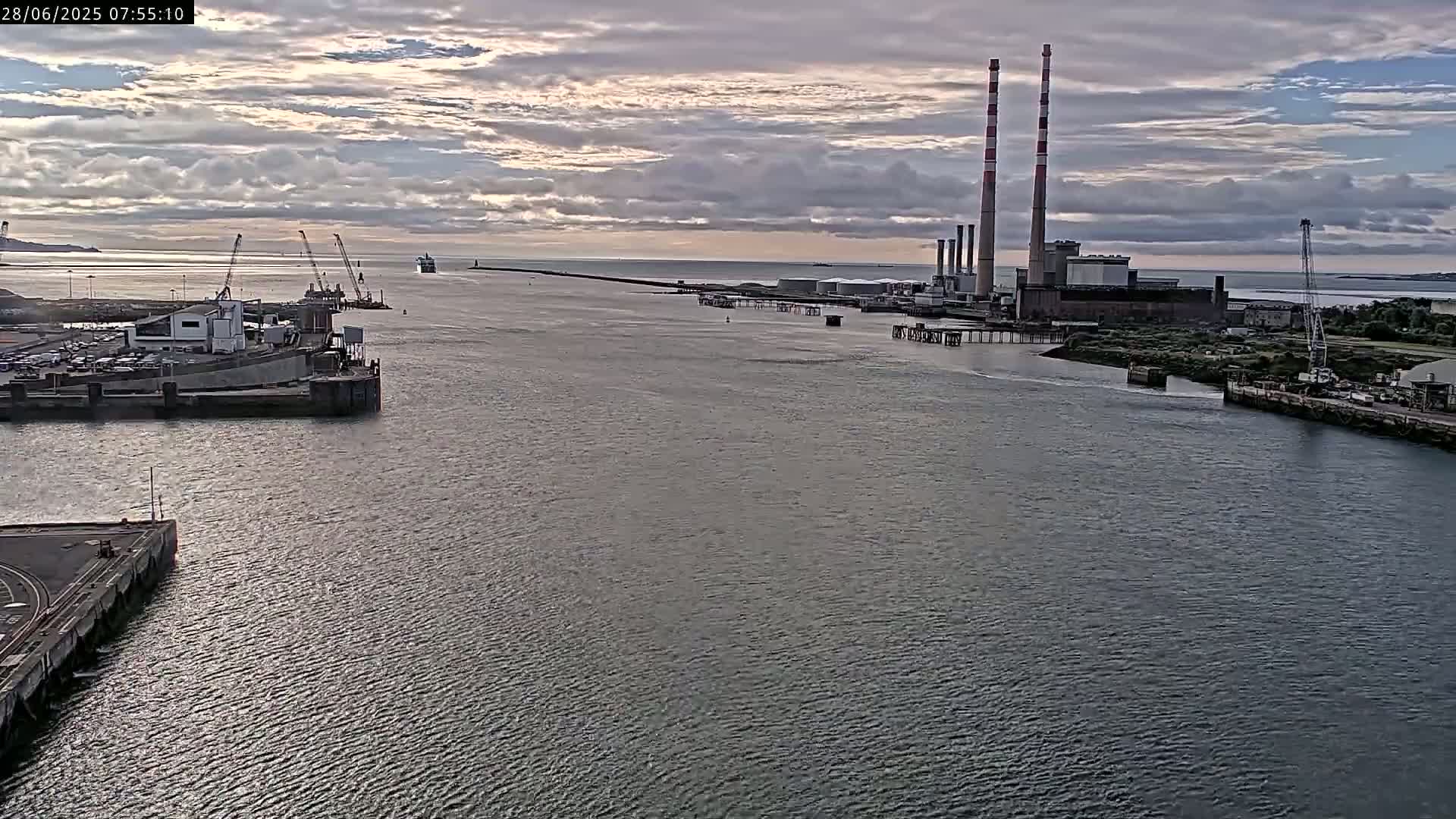 A large body of water shows a port with docked ships and a power plant with tall smokestacks under a partly cloudy sky.