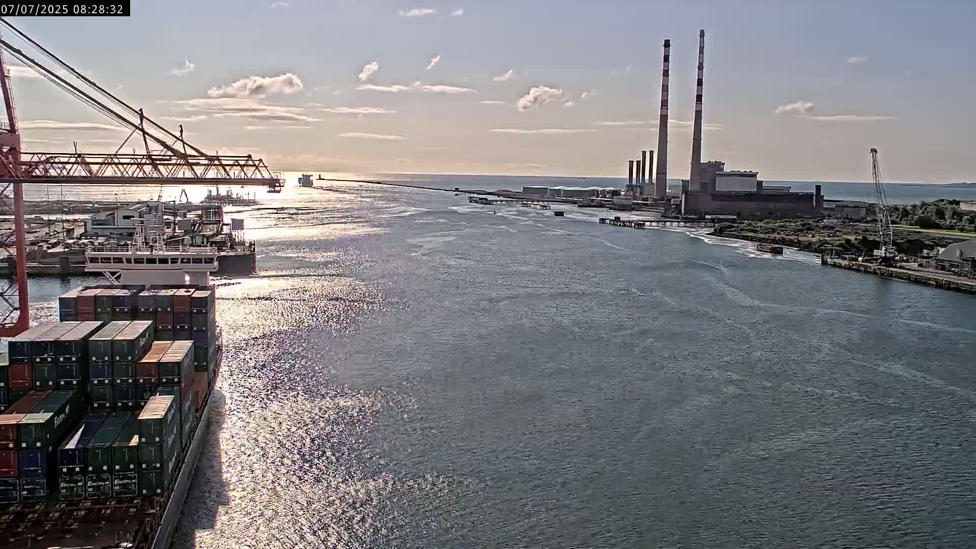 A container ship sails past a port with a large industrial plant and tall chimneys under a partly cloudy sky.