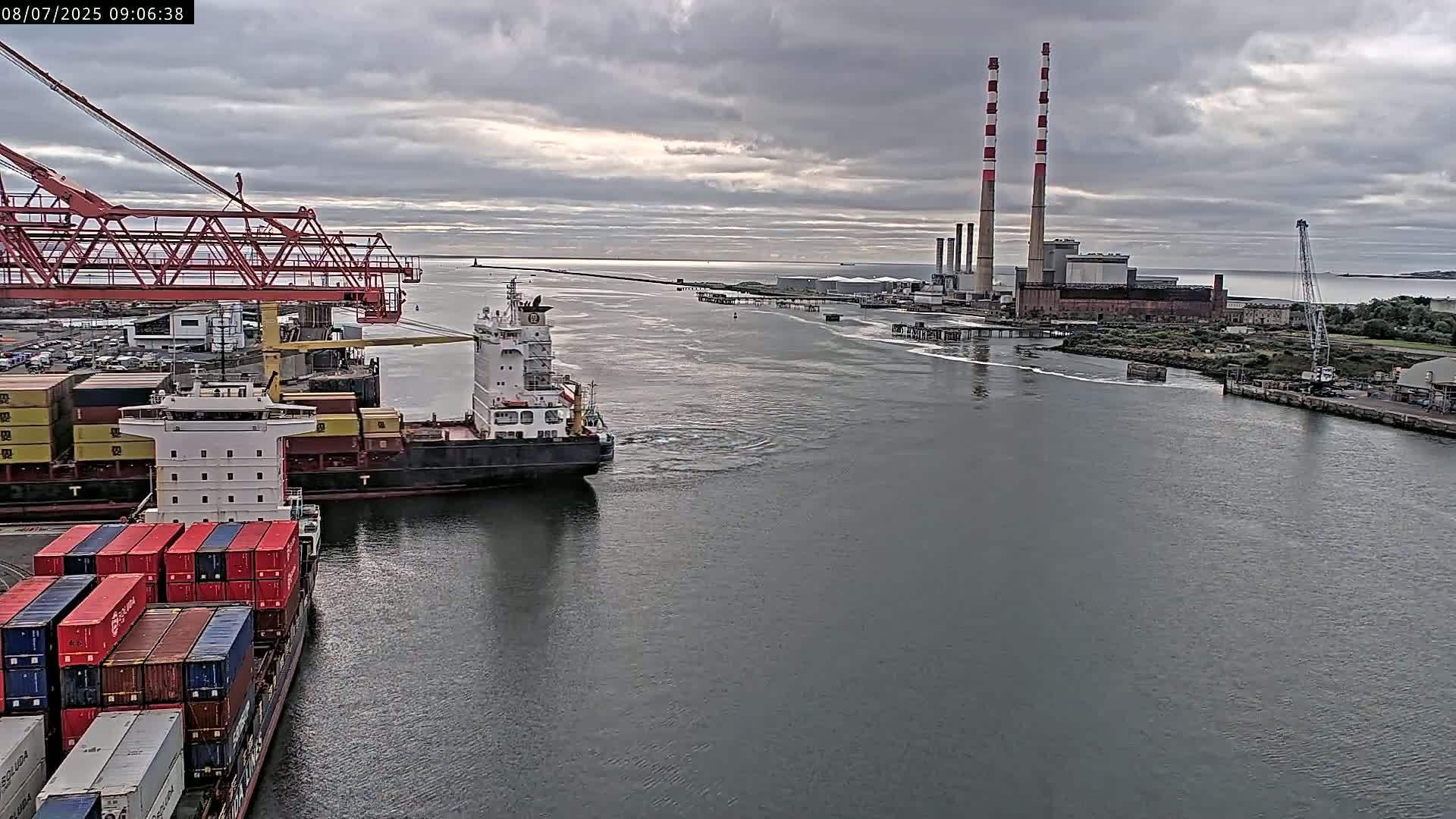 A container ship passes a port under an overcast sky, with two large industrial smokestacks visible in the distance.