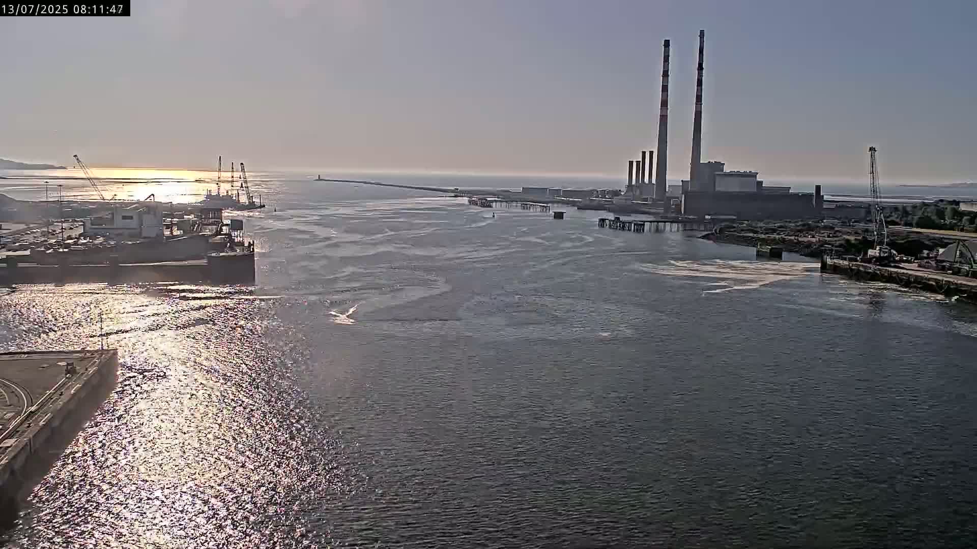 A calm, sunny day reveals a harbor with industrial buildings, docked ships, and a long breakwater extending into the distance.