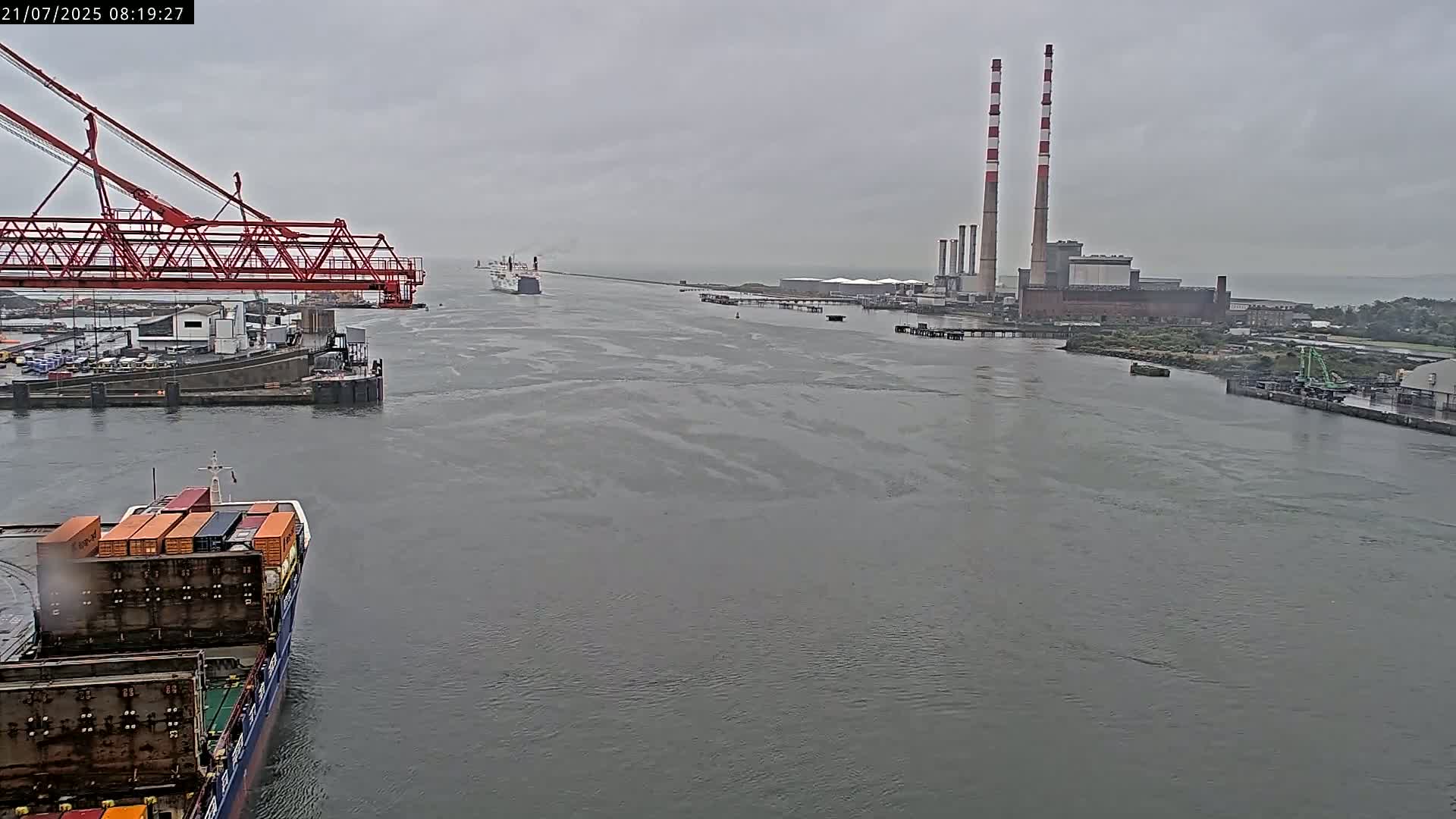 A container ship is in a harbor next to a large industrial facility with tall smokestacks, under an overcast sky on a rainy day.