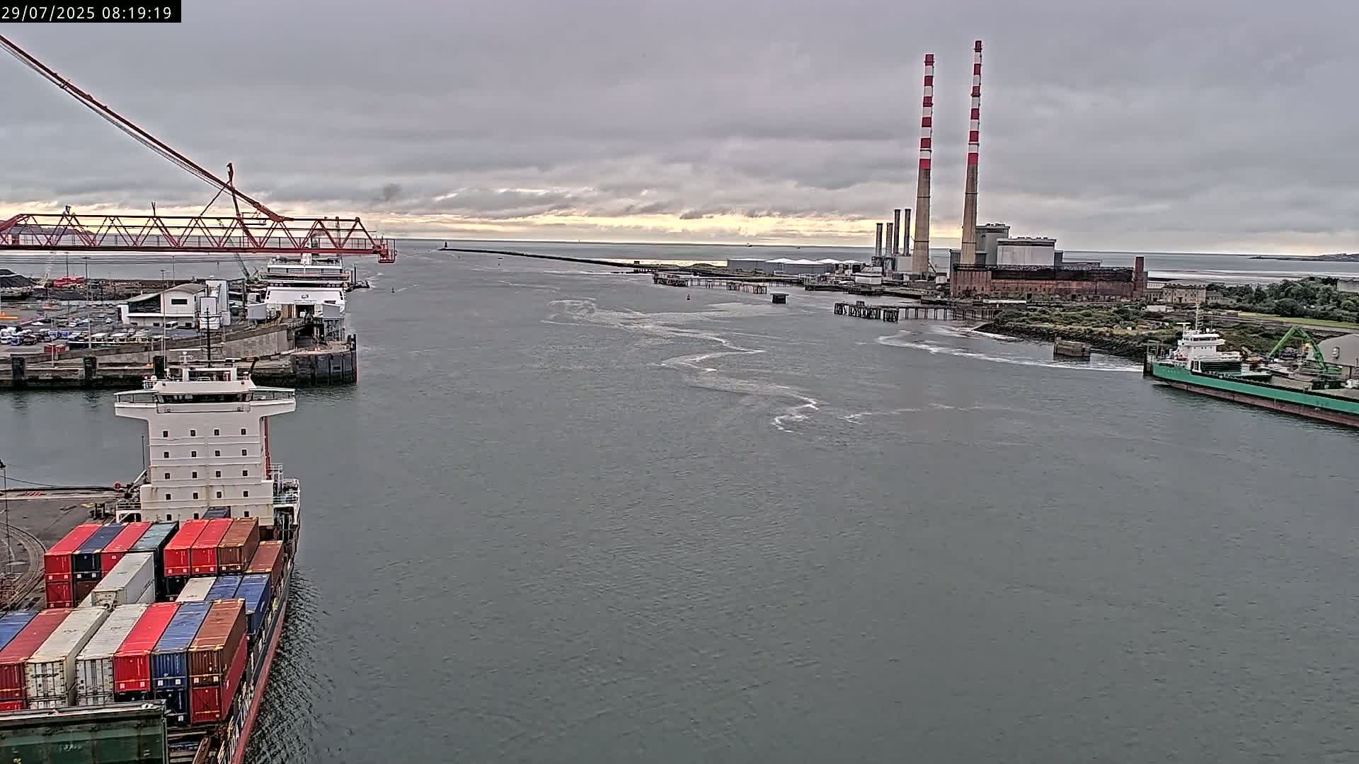 A container ship is docked at a port under an overcast sky, with a ferry in the background near a power plant and a red bridge.