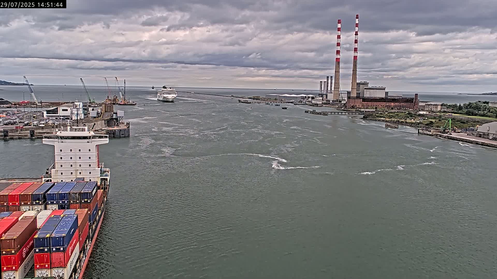A container ship is in a harbor under a cloudy sky, with a large cruise ship and a power plant visible in the distance.