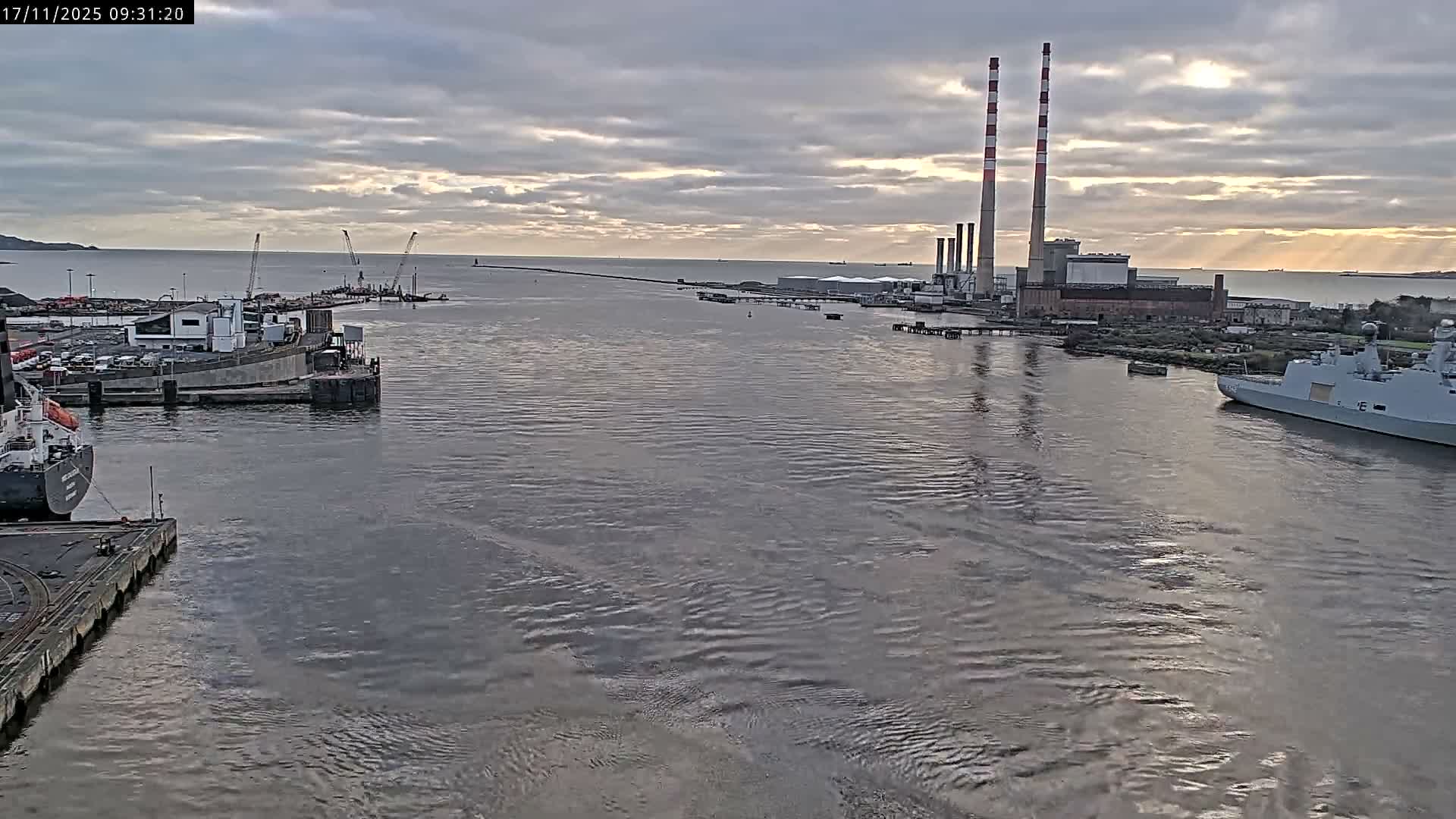 An industrial port and waterway are viewed under an overcast sky with hints of sunlight, featuring prominent red-and-white striped smokestacks, various buildings, cranes, and multiple ships including a large grey naval vessel.
