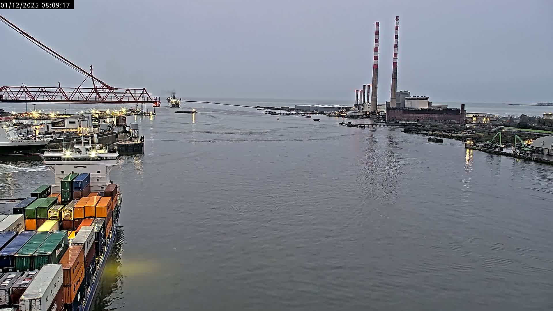 On an overcast early morning, a container ship navigates a busy industrial waterway featuring a large red crane, various port structures, and a power plant with two distinct red and white striped chimneys on the distant shore.