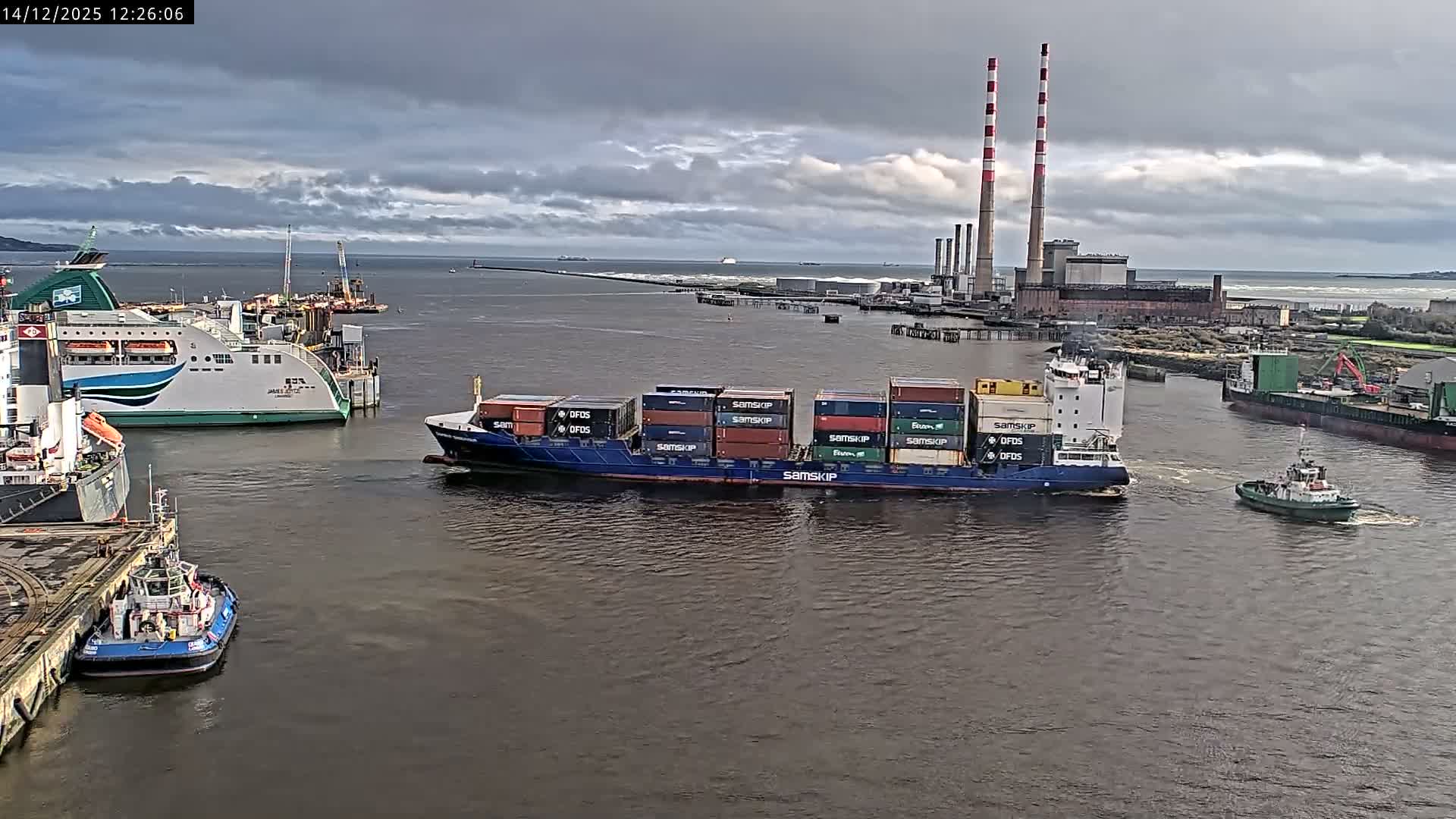 On an overcast early morning, a container ship navigates a busy industrial waterway featuring a large red crane, various port structures, and a power plant with two distinct red and white striped chimneys on the distant shore.