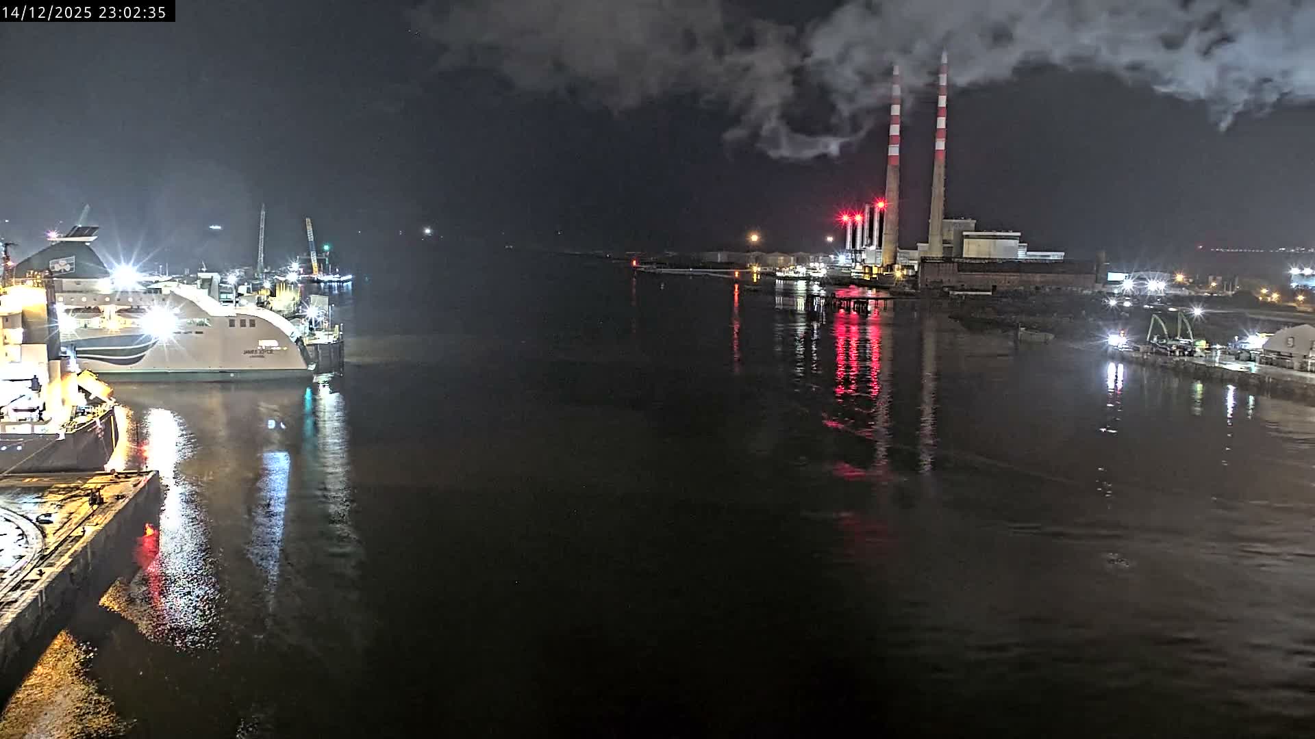 On an overcast early morning, a container ship navigates a busy industrial waterway featuring a large red crane, various port structures, and a power plant with two distinct red and white striped chimneys on the distant shore.
