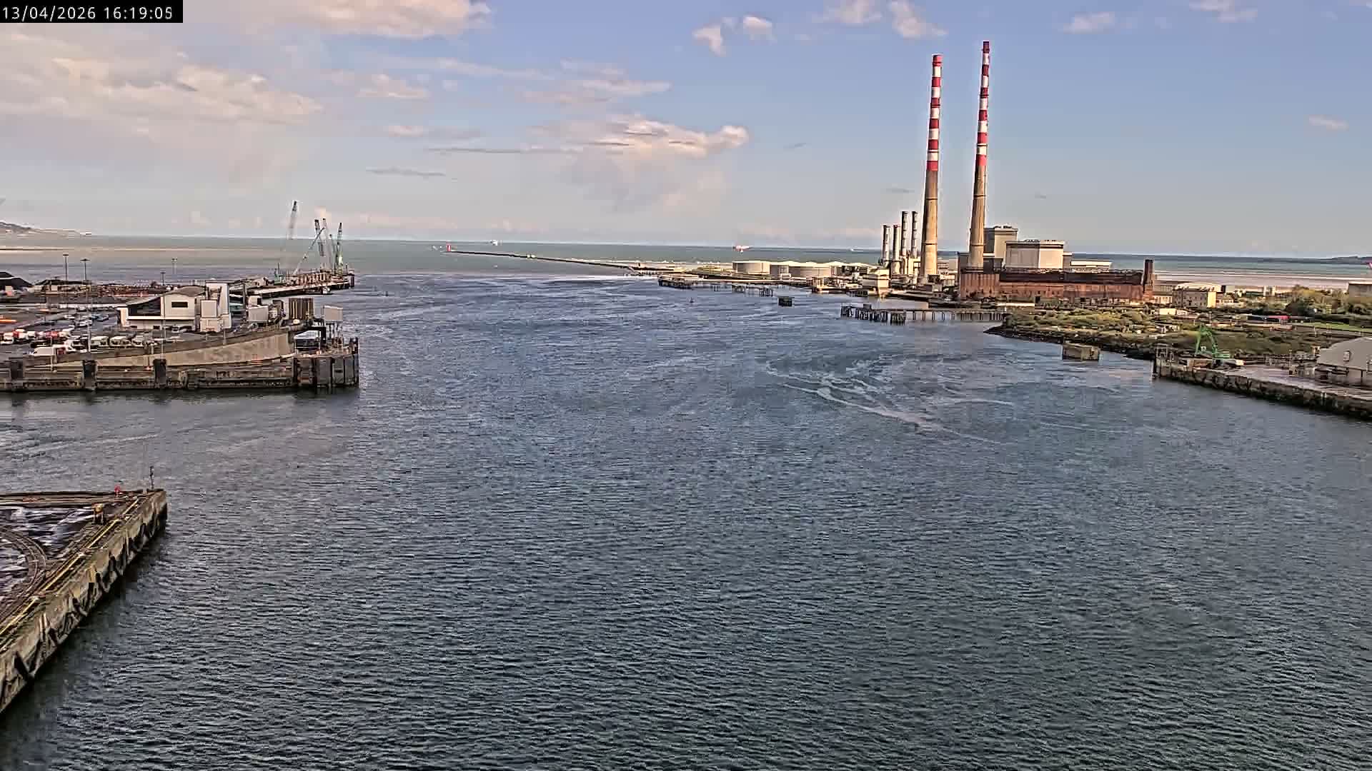 On an overcast early morning, a container ship navigates a busy industrial waterway featuring a large red crane, various port structures, and a power plant with two distinct red and white striped chimneys on the distant shore.