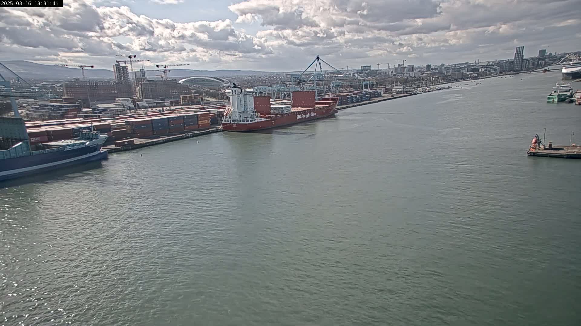 A large container ship is docked at a busy port under a partly cloudy sky.