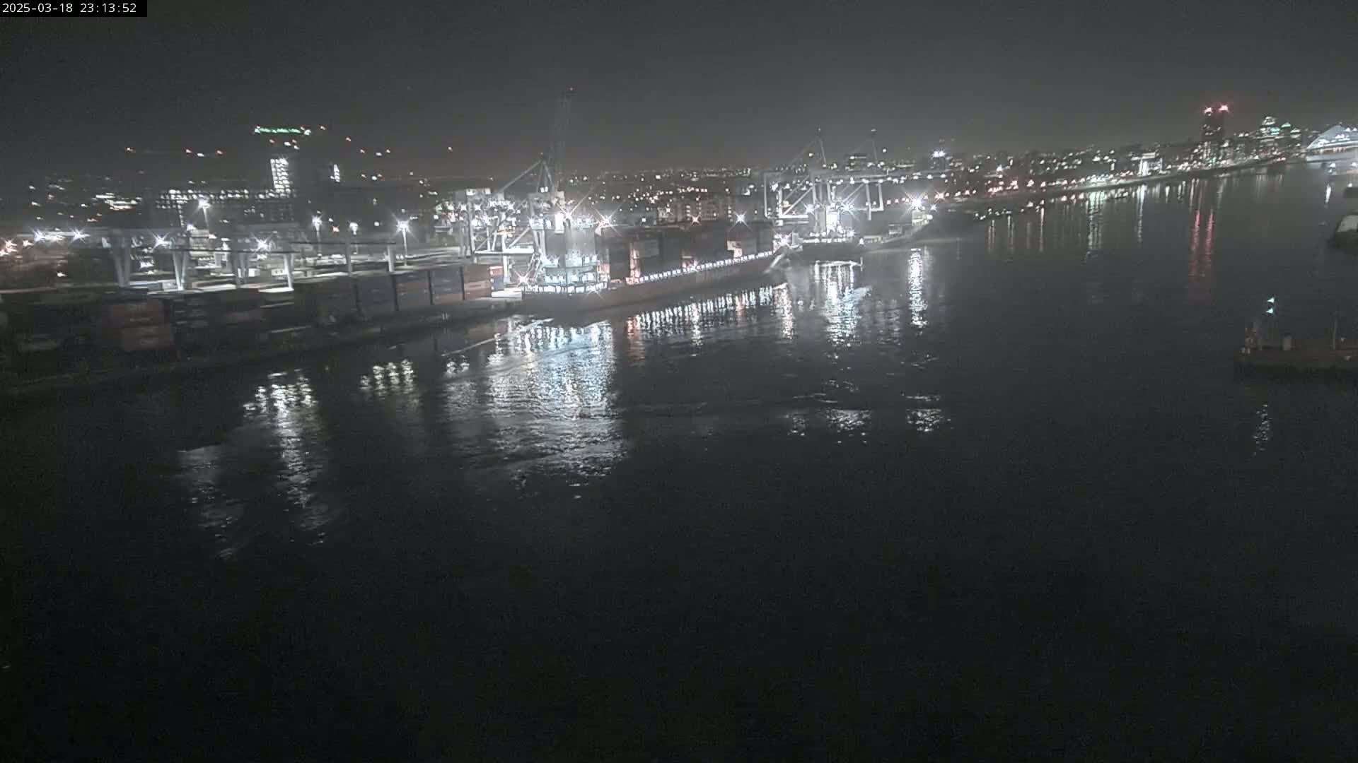 A large cargo ship is docked at a nighttime port, with calm water reflecting the city lights in the background.