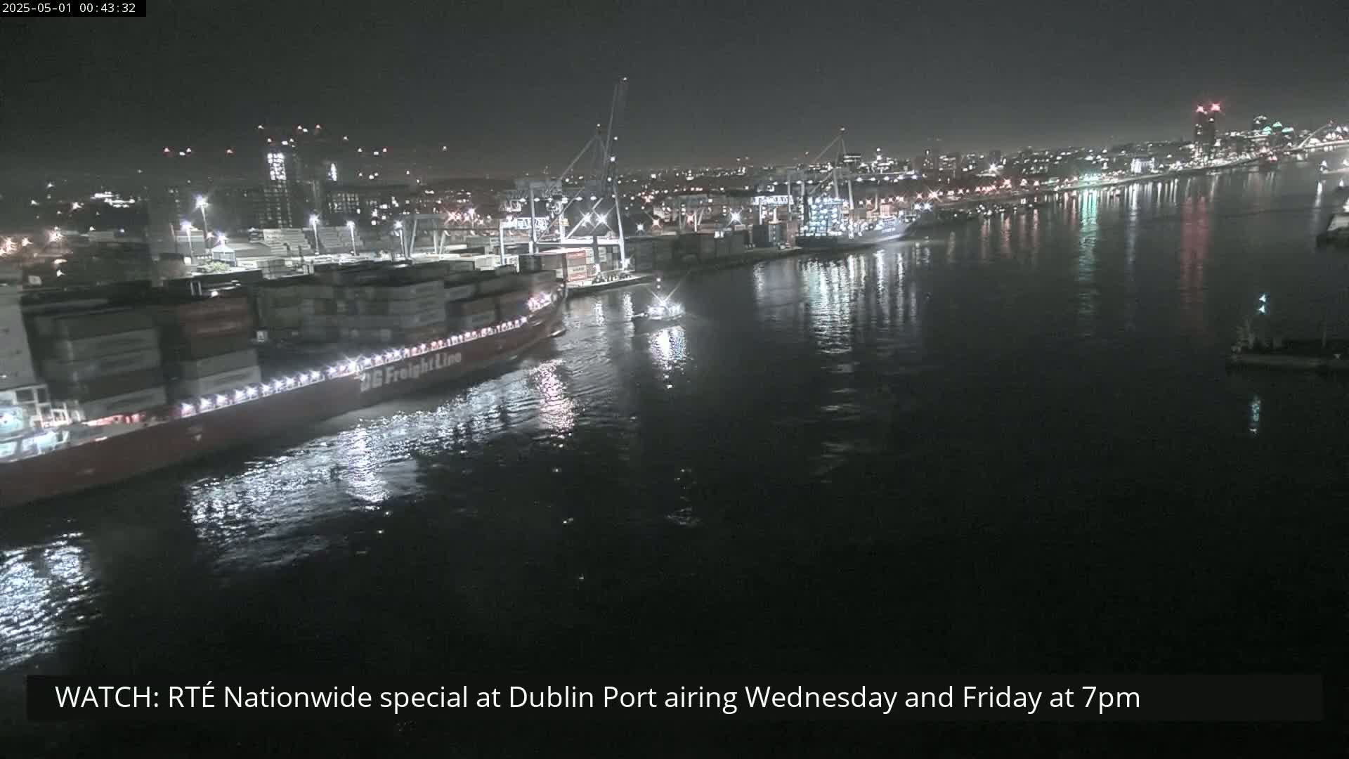 A large container ship is moored at a busy port at night under a clear, dark sky.