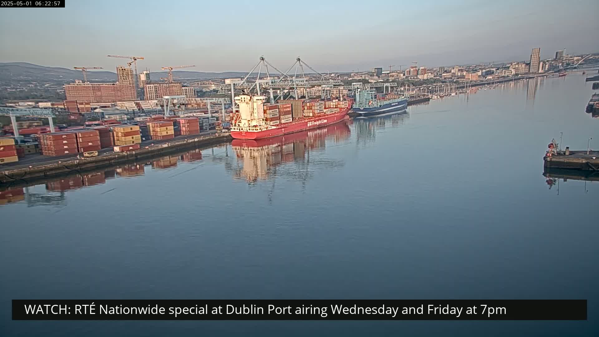 A large red container ship is docked at a port, with numerous other ships and containers visible under a clear, calm sky.