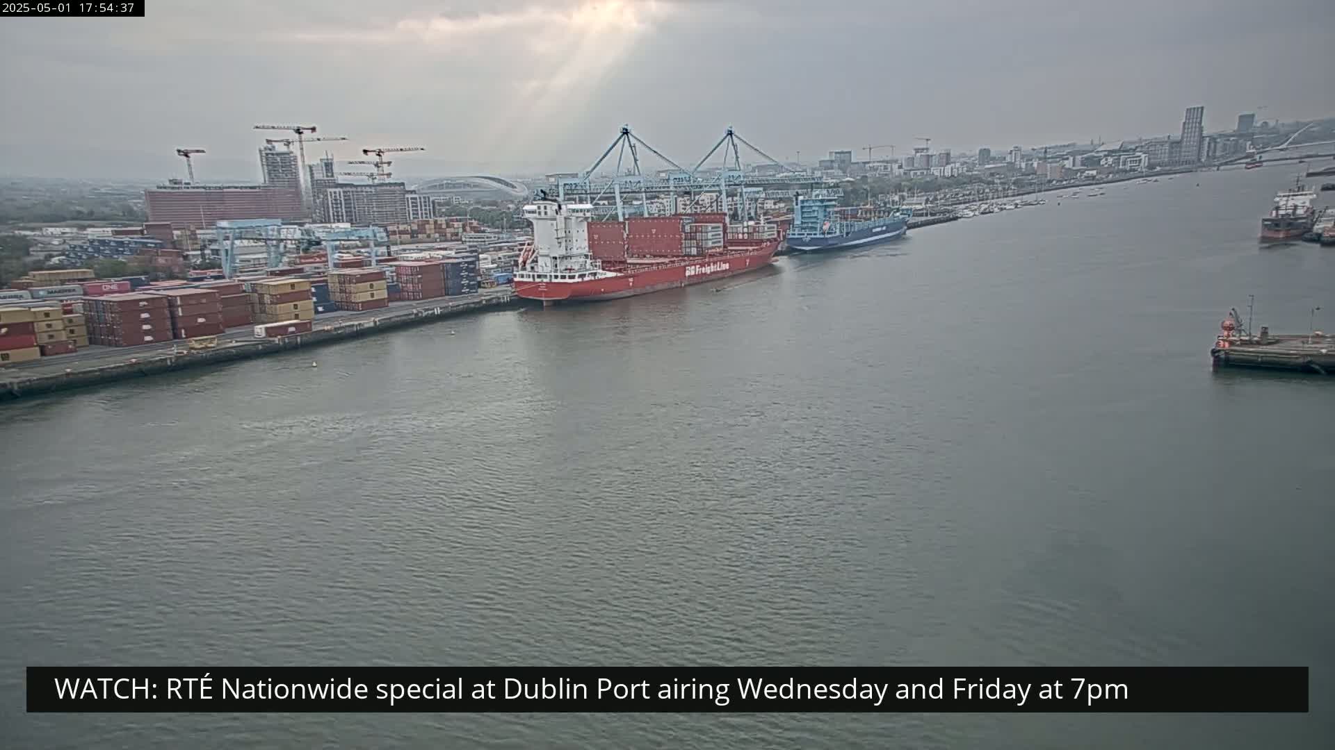 A large red container ship and a smaller blue ship are docked at a port, under an overcast sky.