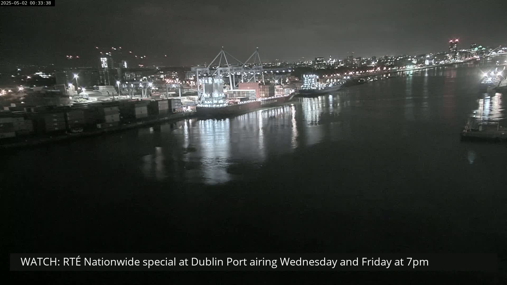 A nighttime view of a harbor with a large cargo ship docked and city lights in the background under a dark sky.