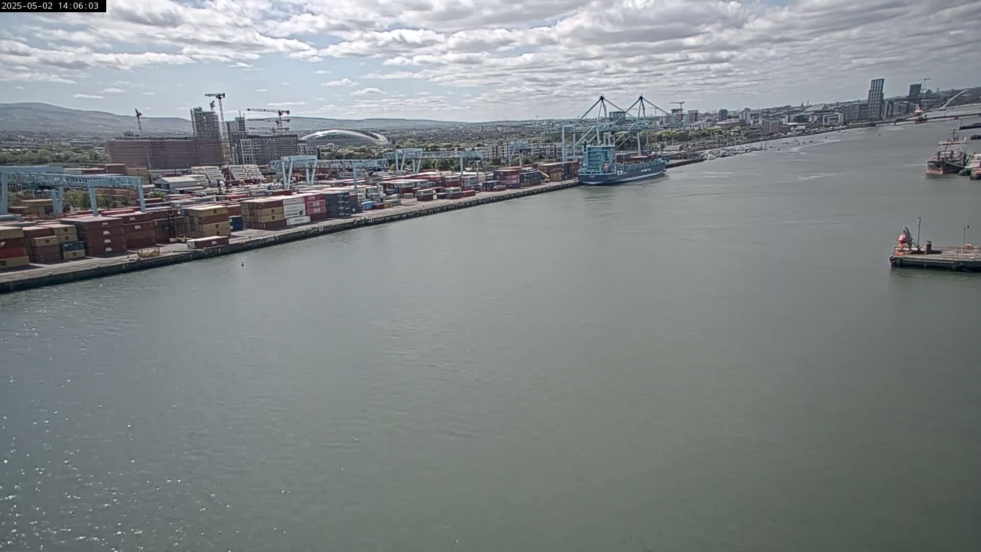 A container ship is docked at a port under a partly cloudy sky, with numerous shipping containers visible along the dock and other vessels in the distance.