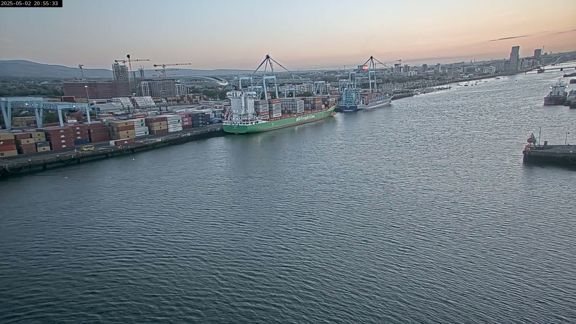 A large container ship and other vessels are docked at a port at twilight, with calm water and a city skyline in the background.
