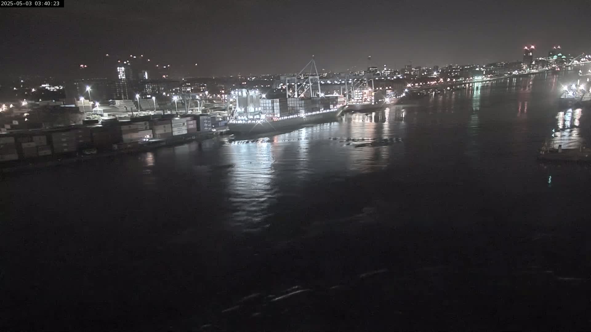A large container ship is docked at a nighttime port, with city lights reflecting on calm water.