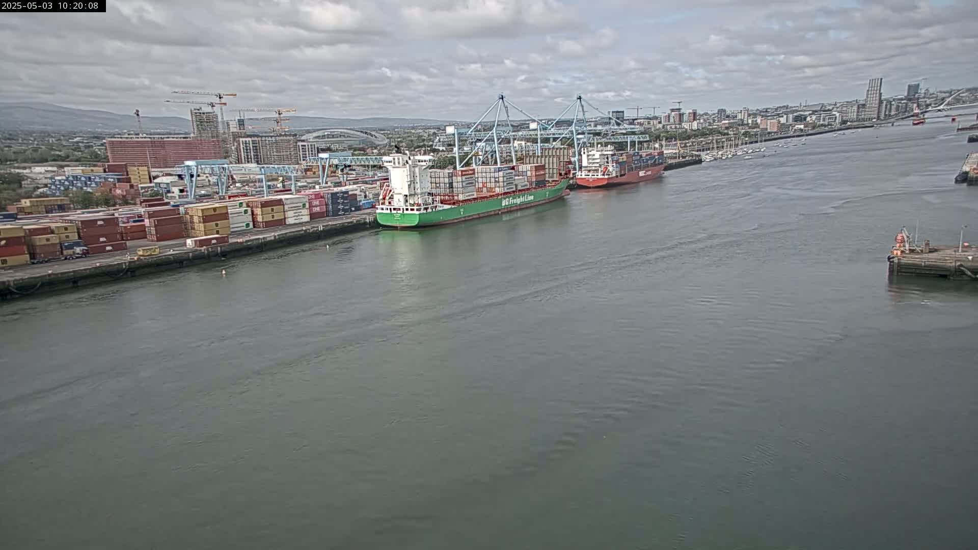 Two container ships are docked at a busy port under a partly cloudy sky.