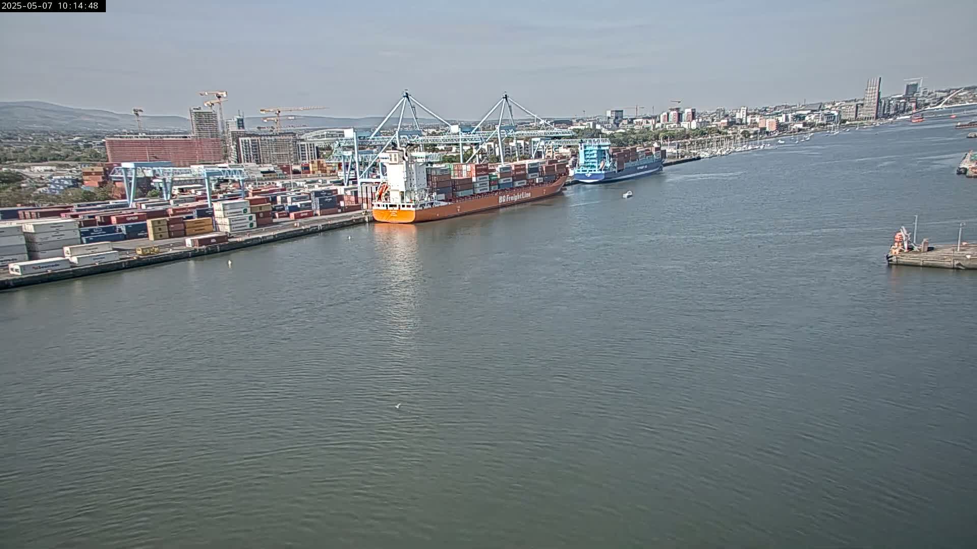 A large container ship and smaller cargo vessel are docked at a port on a calm, sunny day, with a city skyline visible in the background.