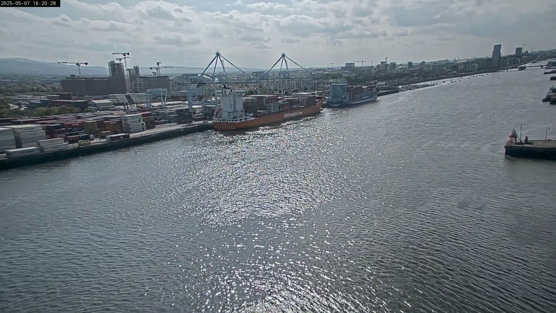 A container ship and other vessels are docked at a port on a partly cloudy day.